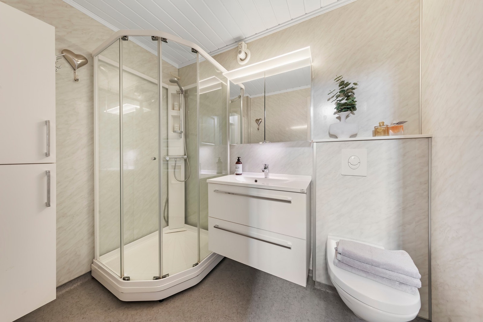 Badet har vinylbelegg med gulvvarme, våtromsplater på vegger og malt panel i innvendig tak. Baderomsinnredning med nedfelt servant, vegghengt toalett og dusjkabinett Galleribilde