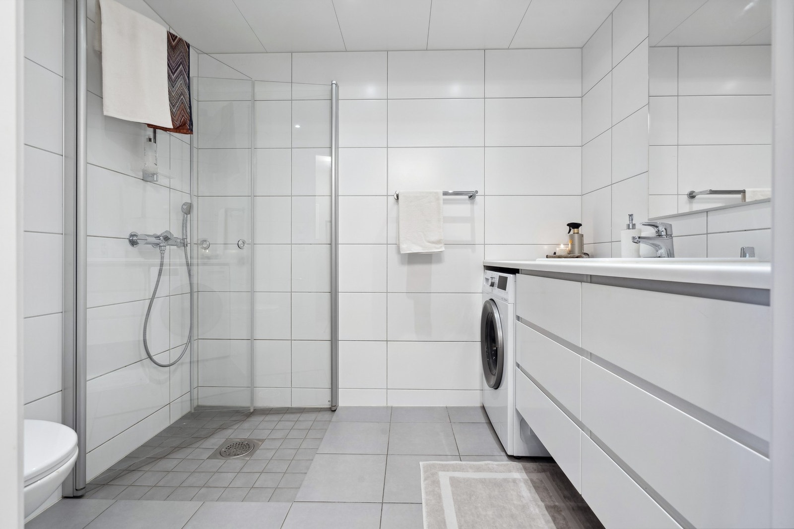 Badet er 4,4 kvm stort og har en tidløs og pen innredning. Dusjdører i glass, kran og avløp for vaskemaskin og veggmontert WC. Galleribilde
