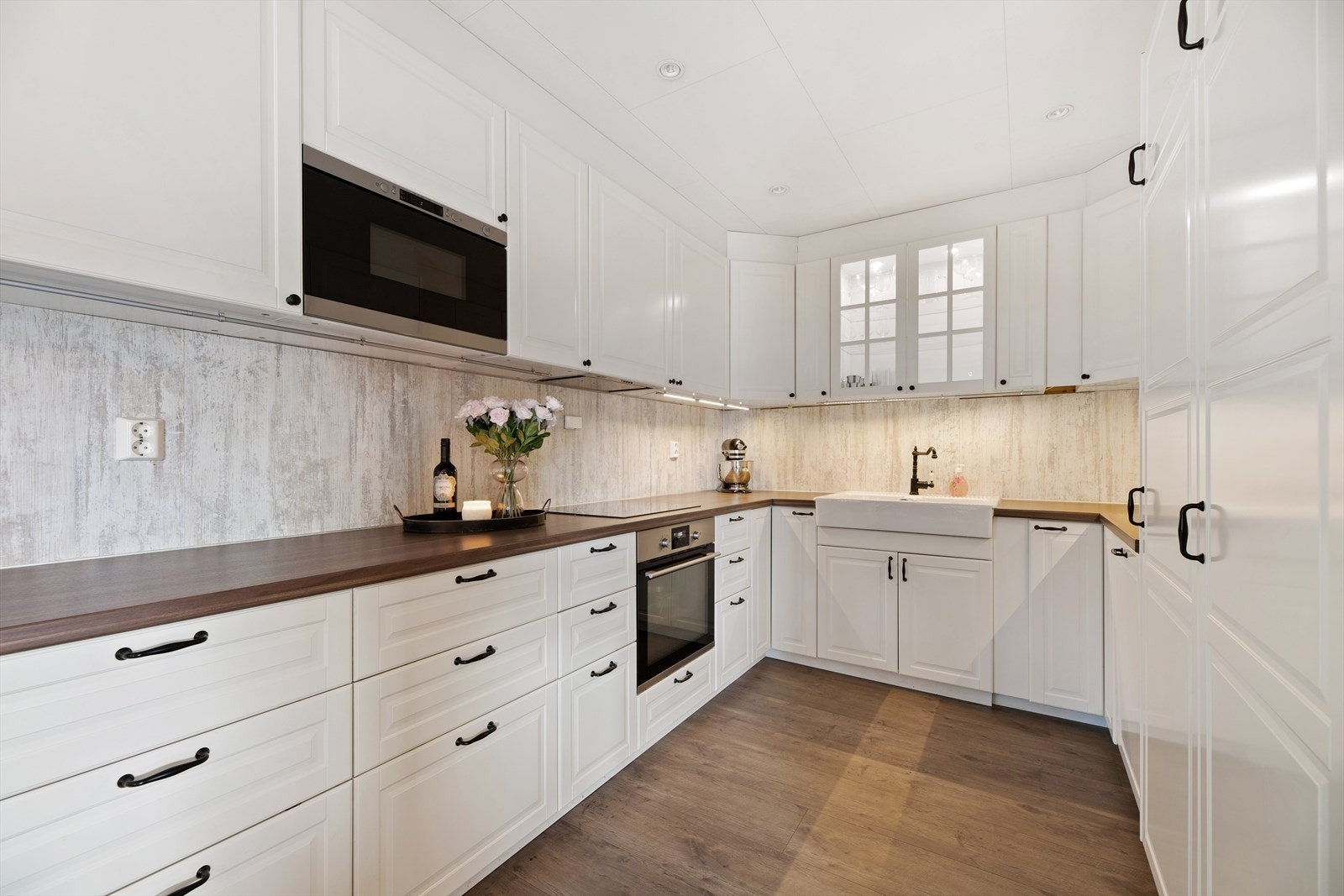 Kjøkkeninnredning med takhøye skap med hvite profilerte fronter, laminat benkeplate og kjøkkenplate på vegg mellom benkeplate og overskap. Galleribilde