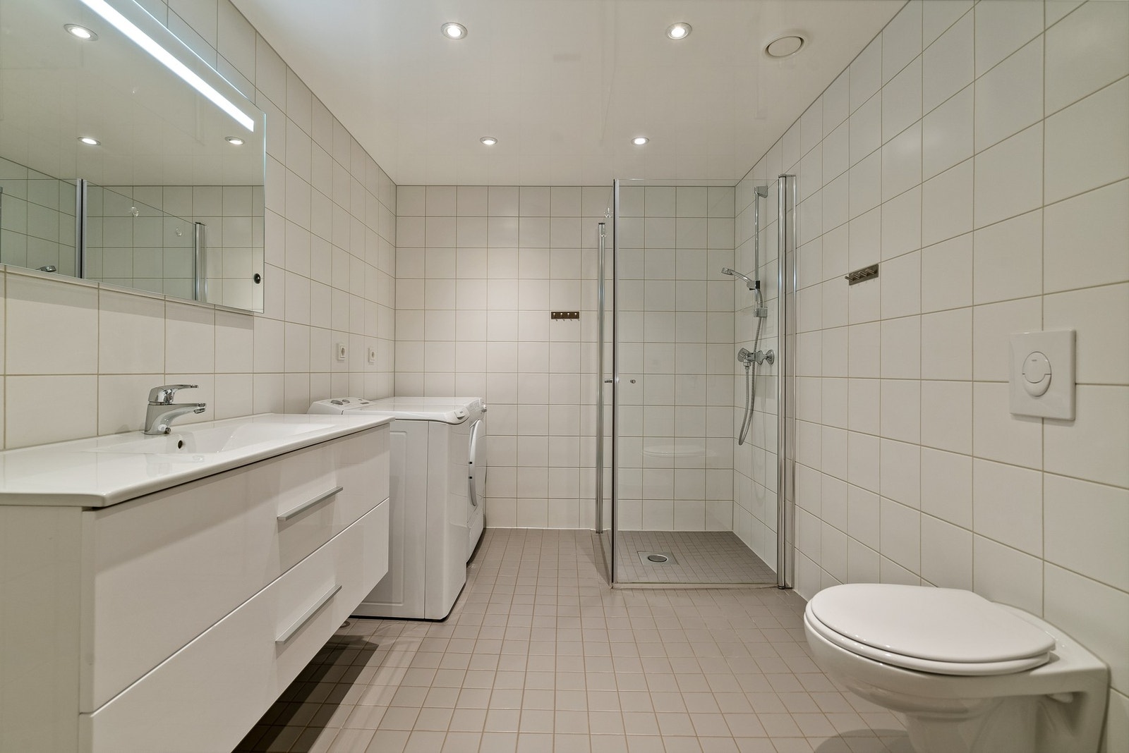 Lyst og stilrent bad med dusjnisje med glassdører, servant, toalett og opplegg til vaskemaskin. Galleribilde