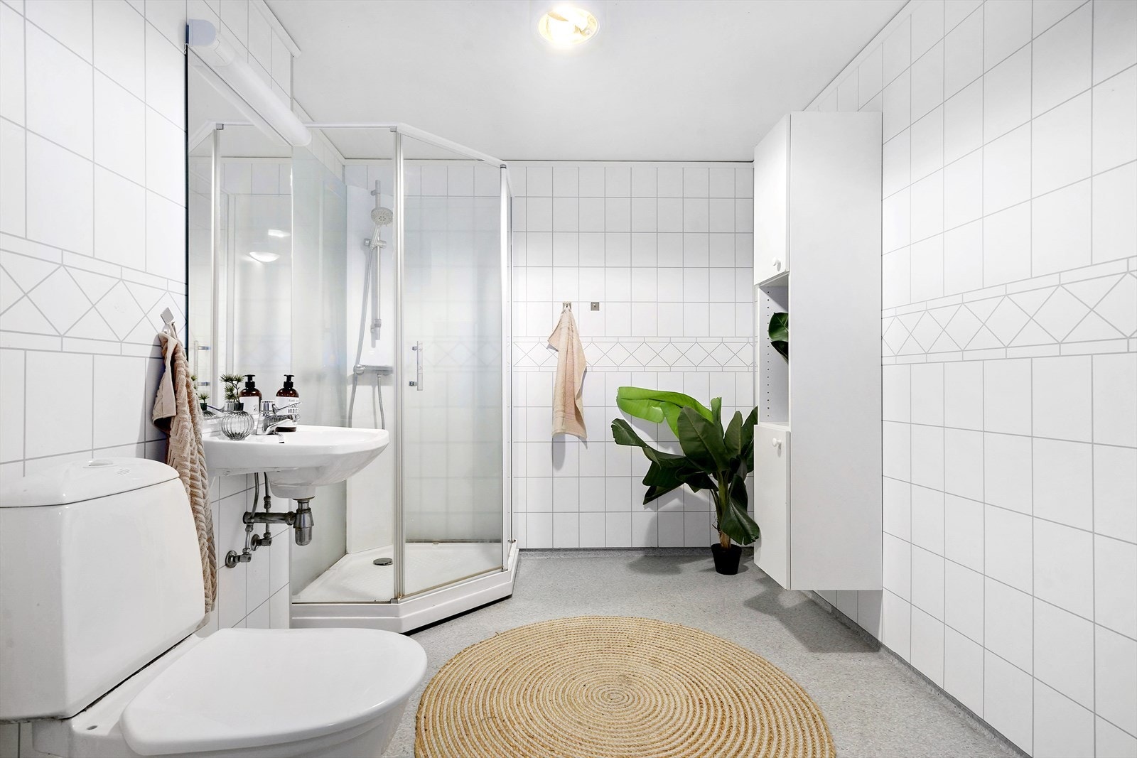 Lyst og pent bad med heldekkende dusjkabinett, servant og gulvmontert toalett. Over servanten er det belysningsarmatur. Galleribilde