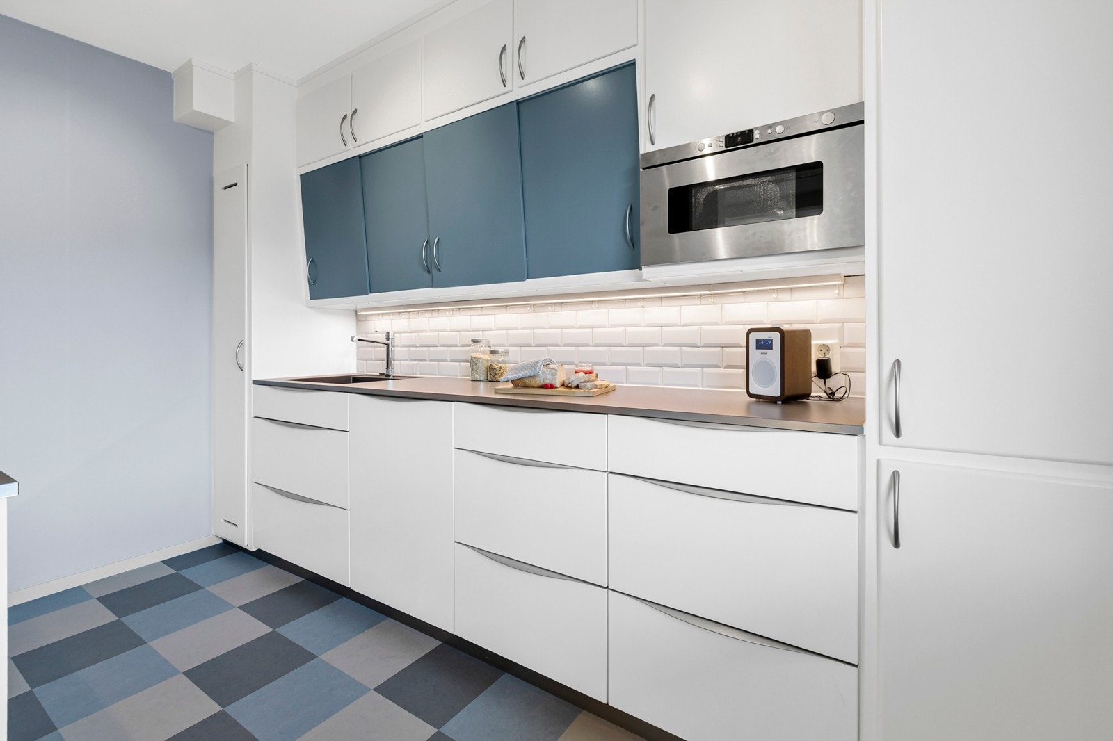 Kjøkkeninnredning med glatte fronter. Belysning under overskap. Fliser på vegg over benkeskap. Laminat benkeplate med planlimt kum i rustfritt stål. Kullfilterventilator. Galleribilde