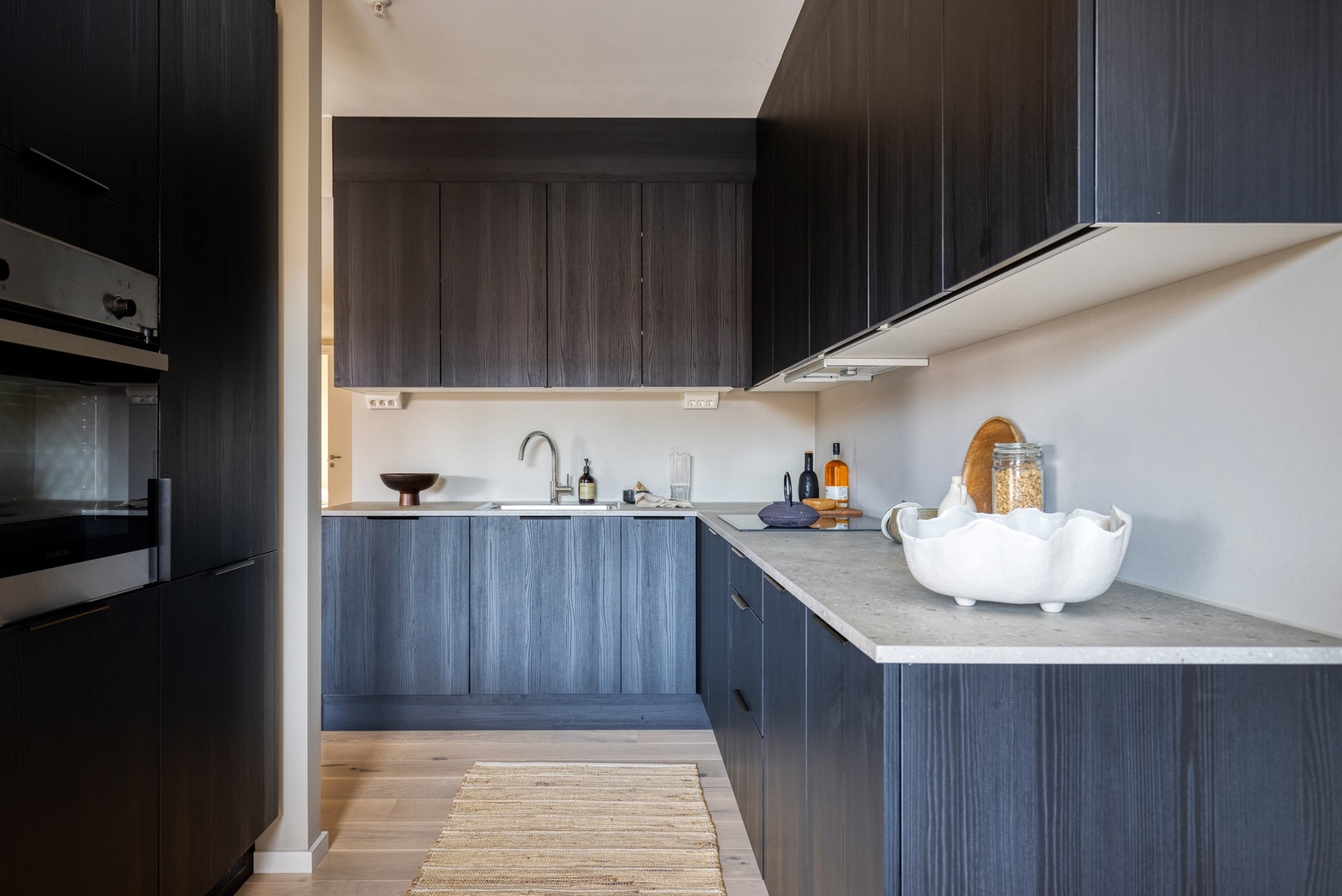 Lekkert HTH-eikekjøkken i mørk, glatt utførelse med laminat benkeplate, utslagsvask med ett-greps blandebatteri samt stikkontakter og belysning under overskap. Galleribilde