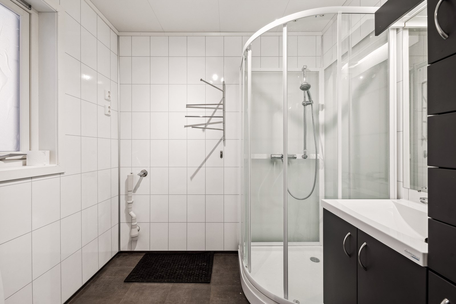 Baderom med baderomsplater på vegger. Rommet er innredet med heldekkende servant over en innredning med skuffer og speil, toalett, dusjkabinett, opplegg for vaskemaskin, avtrekksvifte og gulvvarme. Galleribilde
