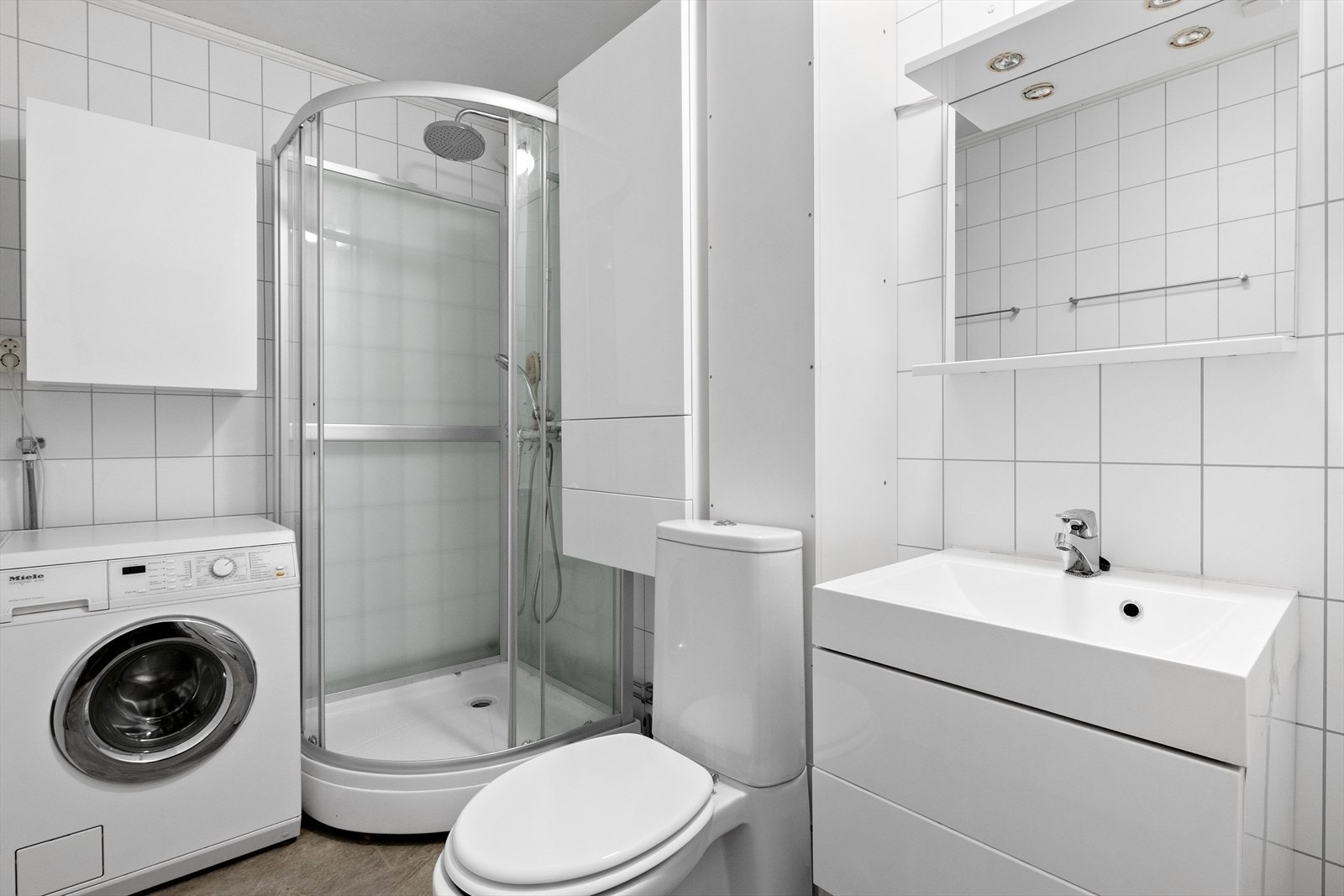 Badet er utstyrt med dusjkabinett, toalett og innredning med slette fronter og nedfelt vask, og veggskap og speil. Det er opplegg for vaskemaskin. Galleribilde