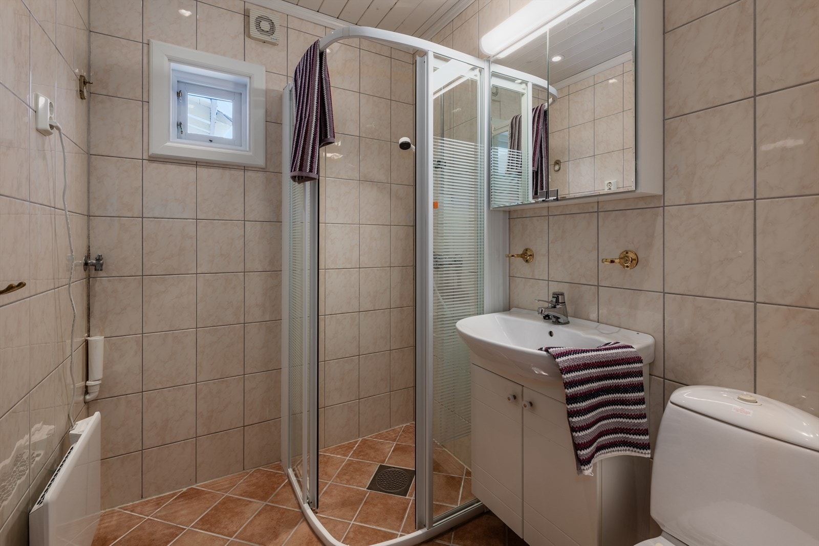 Bad med fliser på gulv og vegger. Dusjvegger i glass, opplegg for vaskemaskin, wc og servant med underskap og speilskap over. Panelovn på vegg. Galleribilde