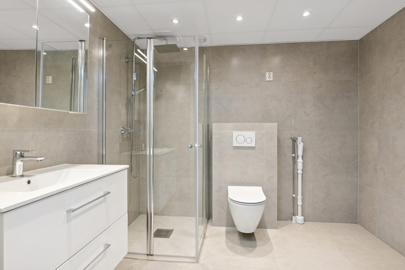 Meget pent flislagt bad med vegghengt klosett, dusjhjørne og servant med innredning. Galleribilde