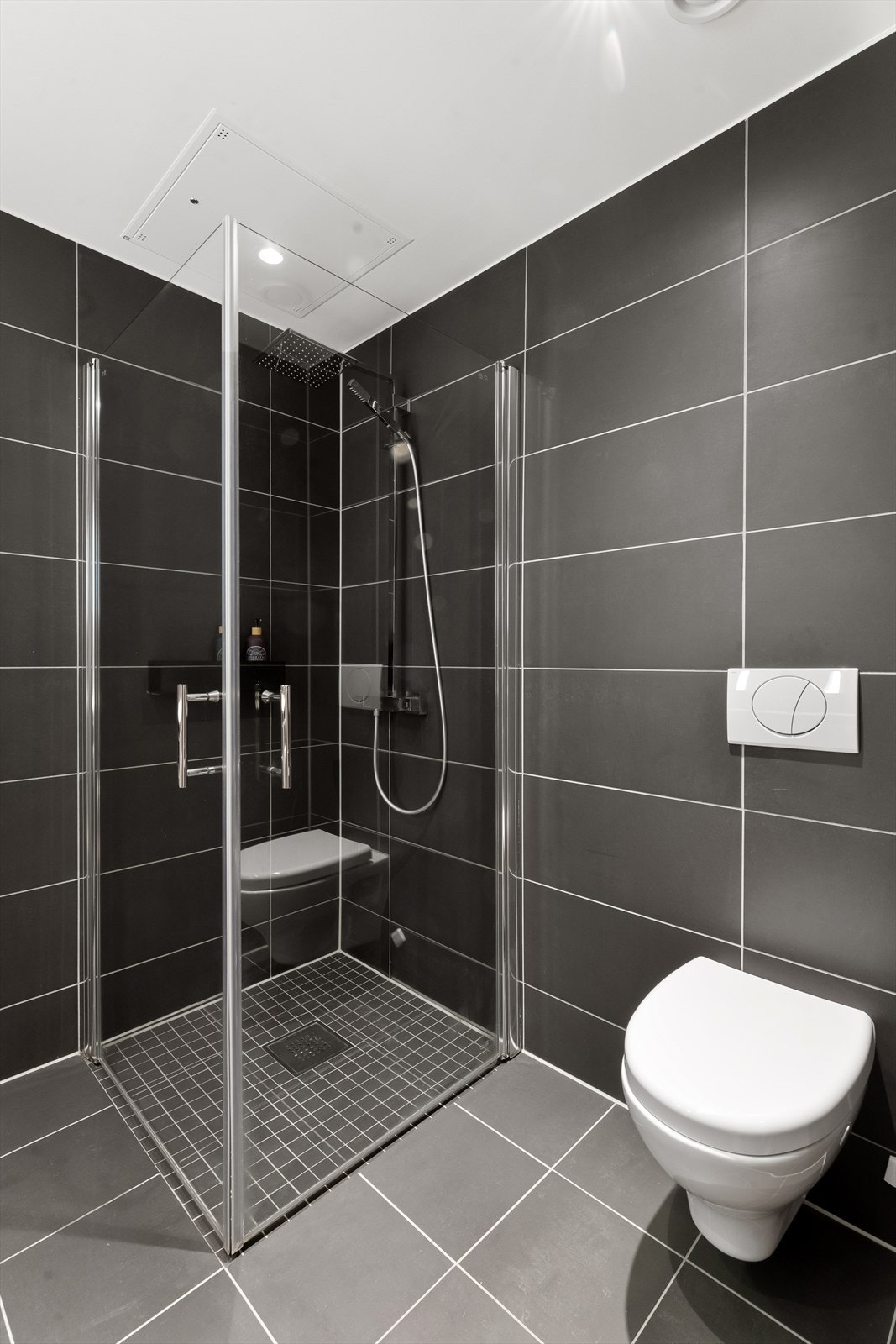 Badet er utstyrt med dusjhjørne med innfellbare dusjdører, veggmontert toalett og baderomsinnredning med servantskap med underlimt servant og speil Galleribilde