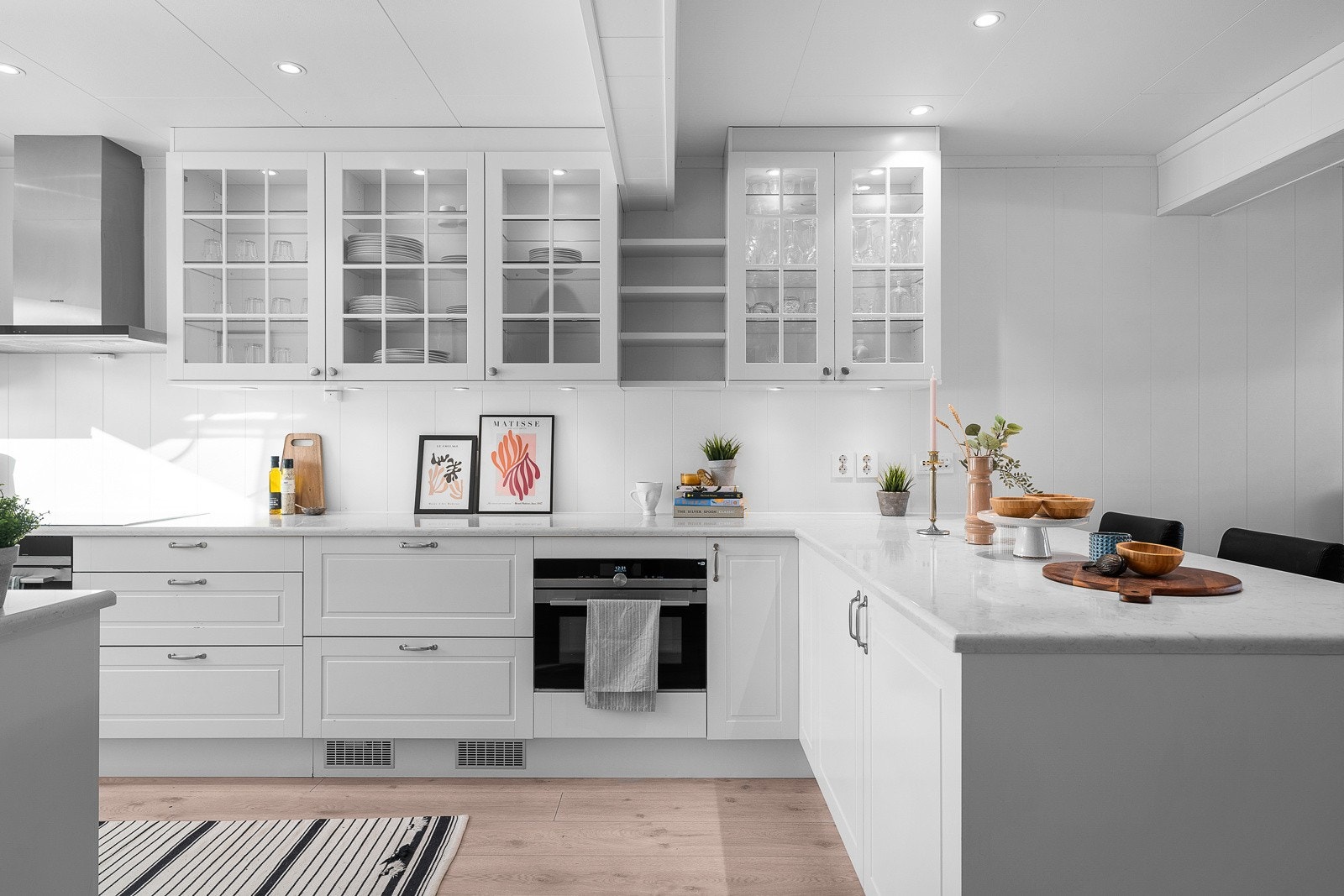 Kjøkkenet har en kledelig, klassisk kjøkkeninnredning i hvitt med mye skap- og benkeplass, samt barløsning mot stuen. Ved siden av den ene komfyren er det to praktiske kjøleskuffer. Galleribilde