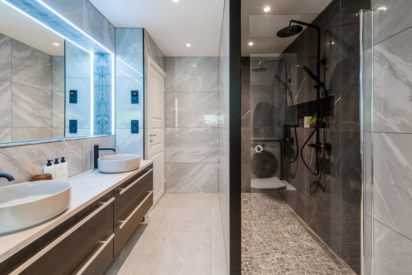 Stort og gjennomført bad med dobbeldusj. Fliser på gulv og vegger. Dobbelservant med dobbel skuffeinnredning under. Innfelt speil på veggen med led belysning. Dobbeldusj med dusjarmaturer i type vannfall. Det er gulvslukrenne mot vegg og glassdør. Galleribilde