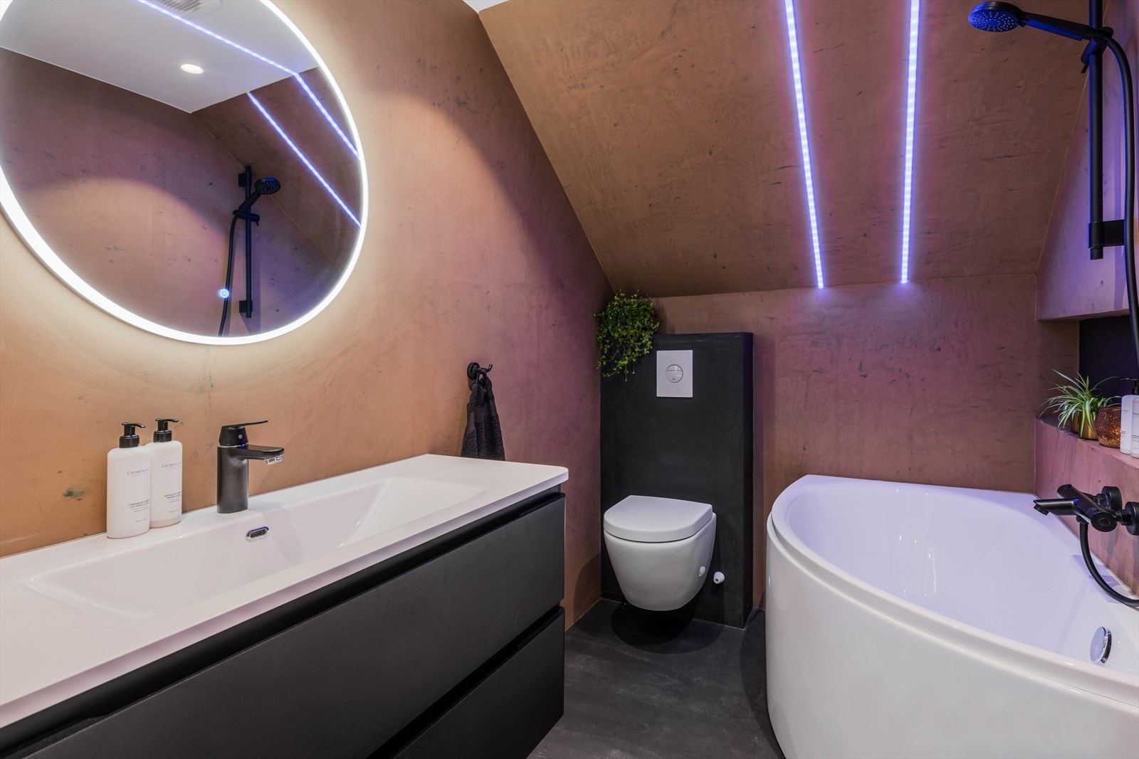 Stilfullt bad med microsement på gulv og vegger. Ledlys i tak og innebygget hylle i vegg. Badekar, vegghengt wc og baderomsinnredning med nedfelt servant med skuffer under. Speil med lys over servant. Galleribilde