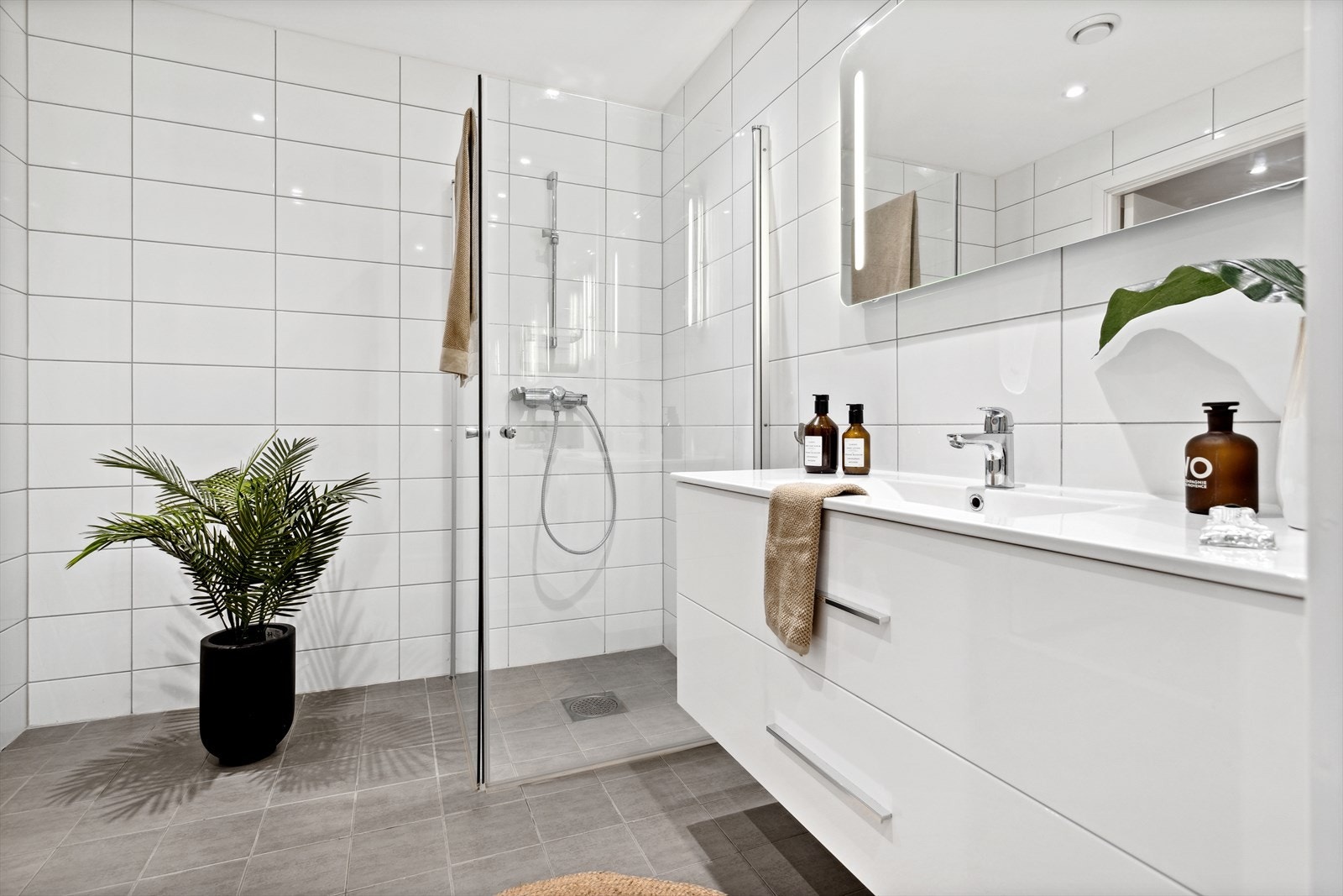 Badet inneholder dusjhjørne med vendbare dusjdører, servant med skuffeseksjon, speil med belysning og vegghengt toalett. Galleribilde