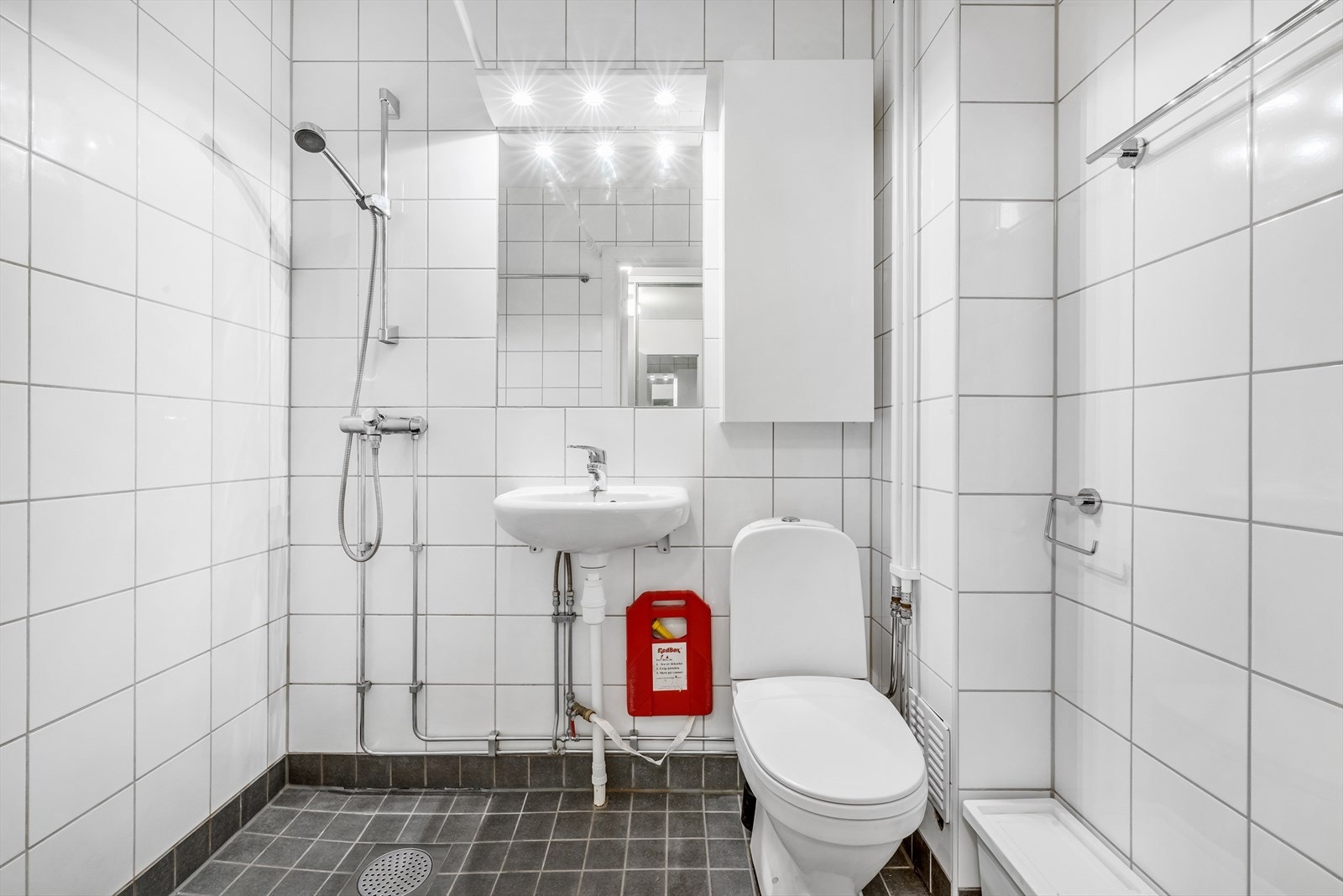 Flislagt bad fra 2016 med innredning bestående av gulvmontert wc, dusjsone og enkel servant med speil, belysning og skap. Galleribilde
