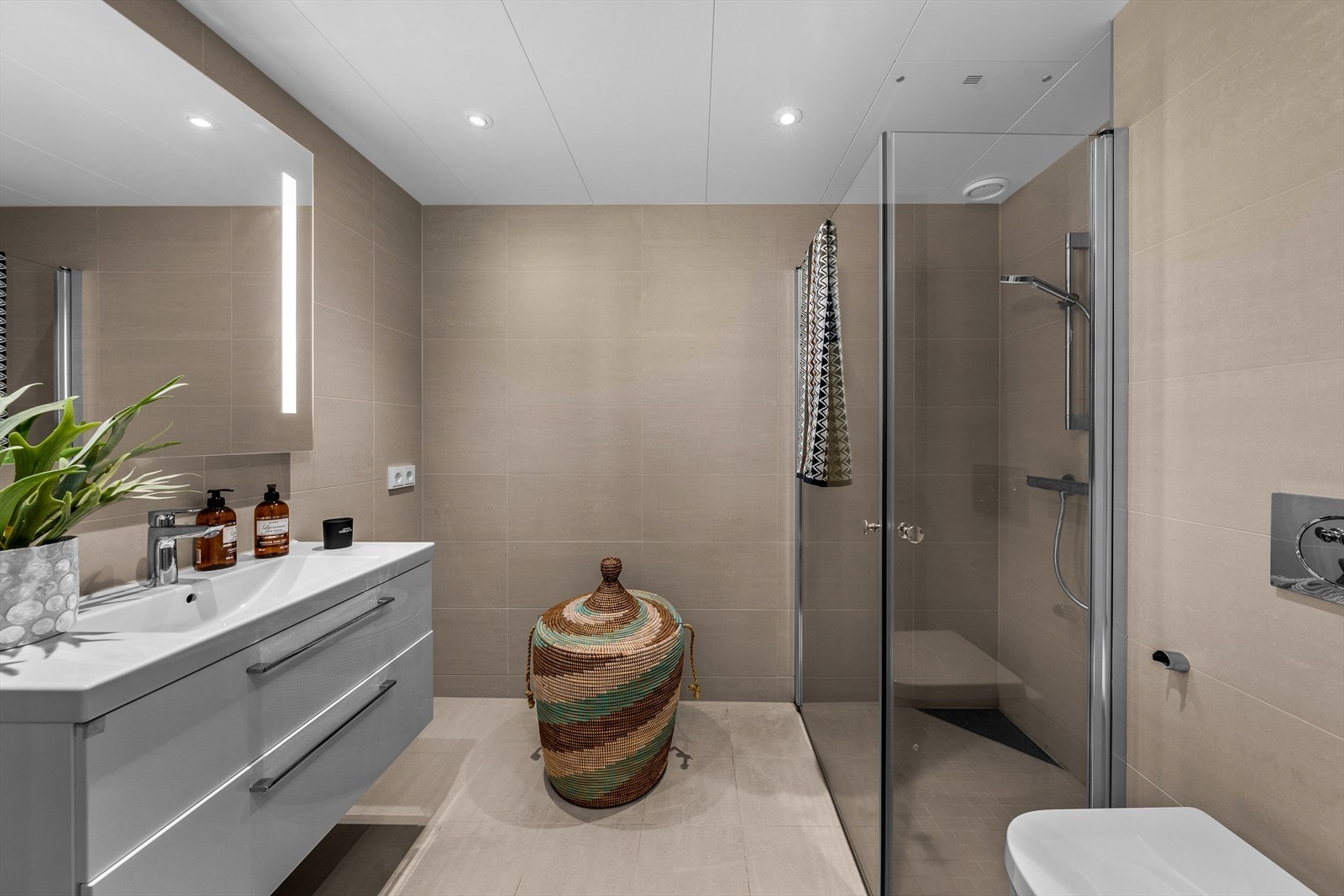 Delikat baderom med flislagte gulv og vegger. Badet er utstyrt med vegghengt toalett, servant med skuffer og dusjnisje med innfellbare glassdører. Galleribilde