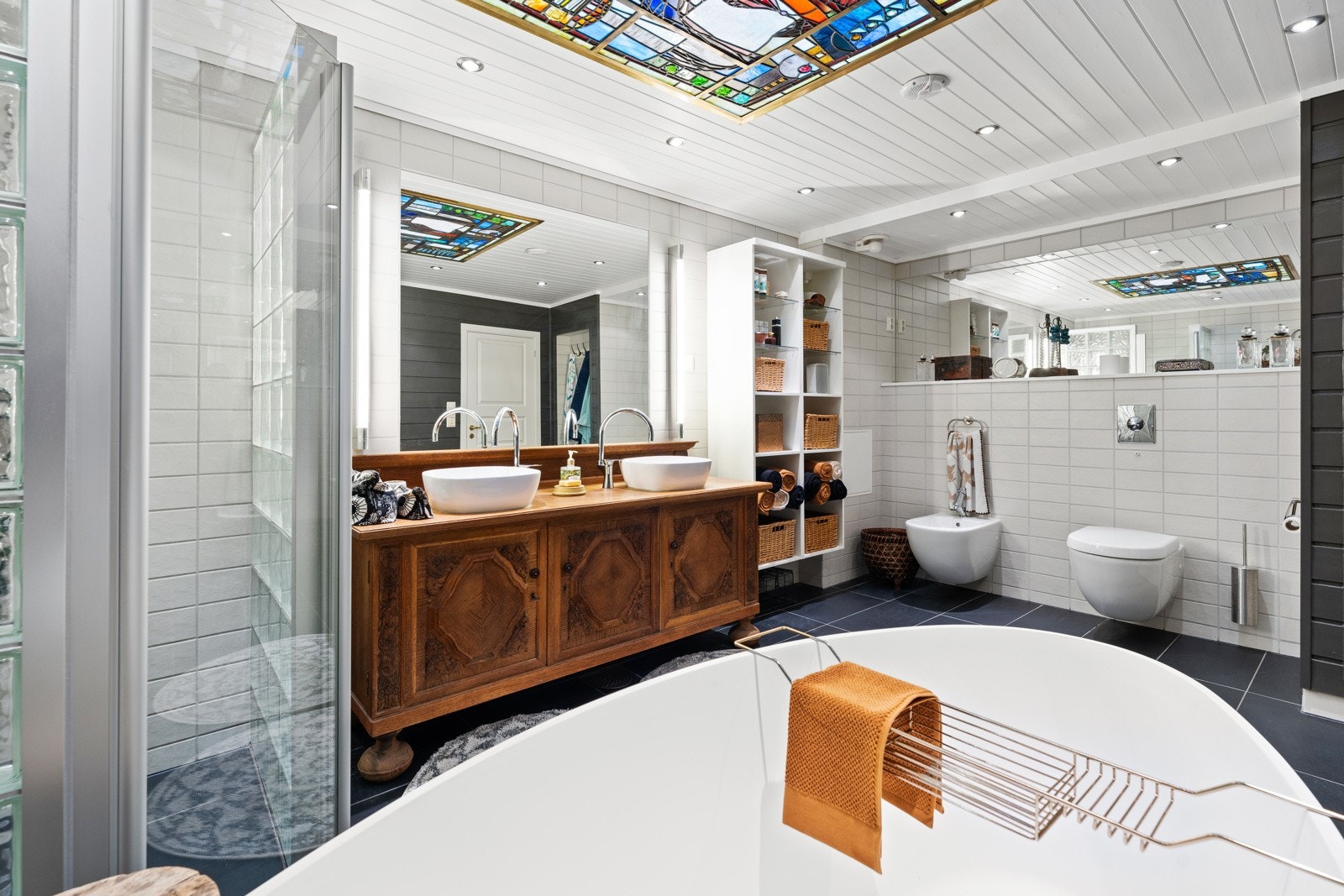 Badet er flislagt på gulv, og med flis/panel på vegger. Romslig dusjhjørne, vegghengt bidé og WC, samt et flott badekar. Legg merke til det stilige glassmaleriet i himlingen med lys. Galleribilde