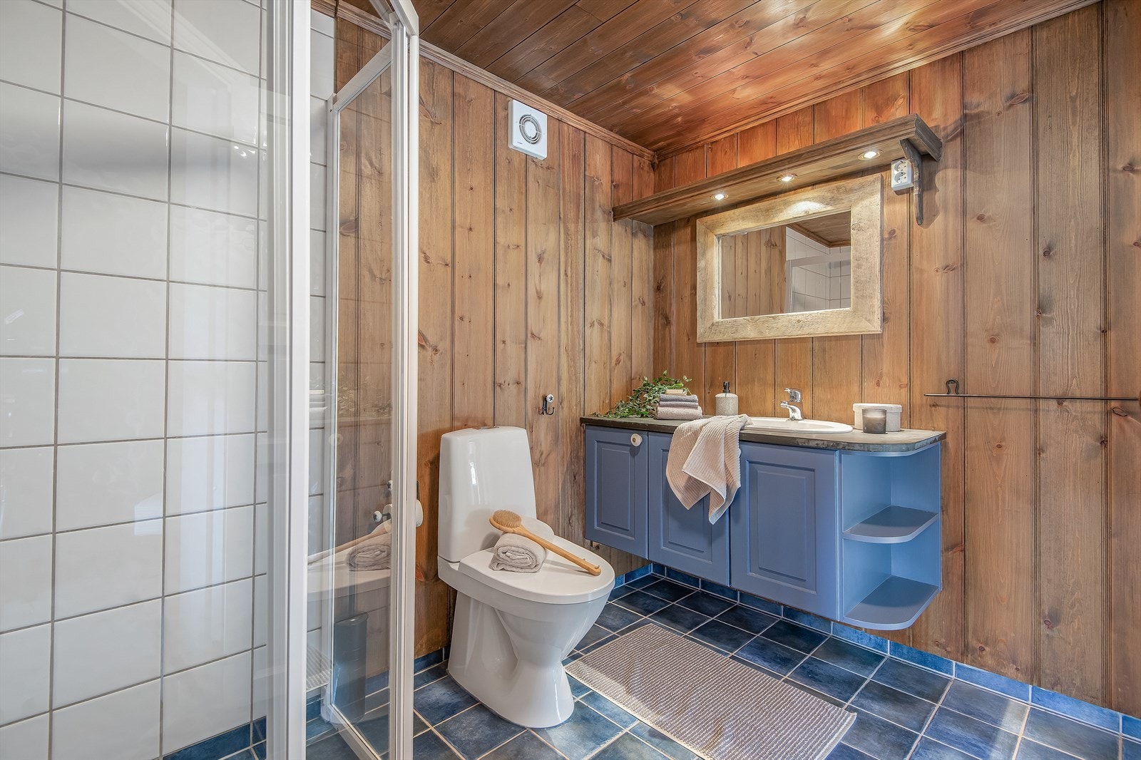 Bad i 2.etasje med blå underskap, speil med tilhørende belysning, dusjvegger og gulvmontert wc. Galleribilde