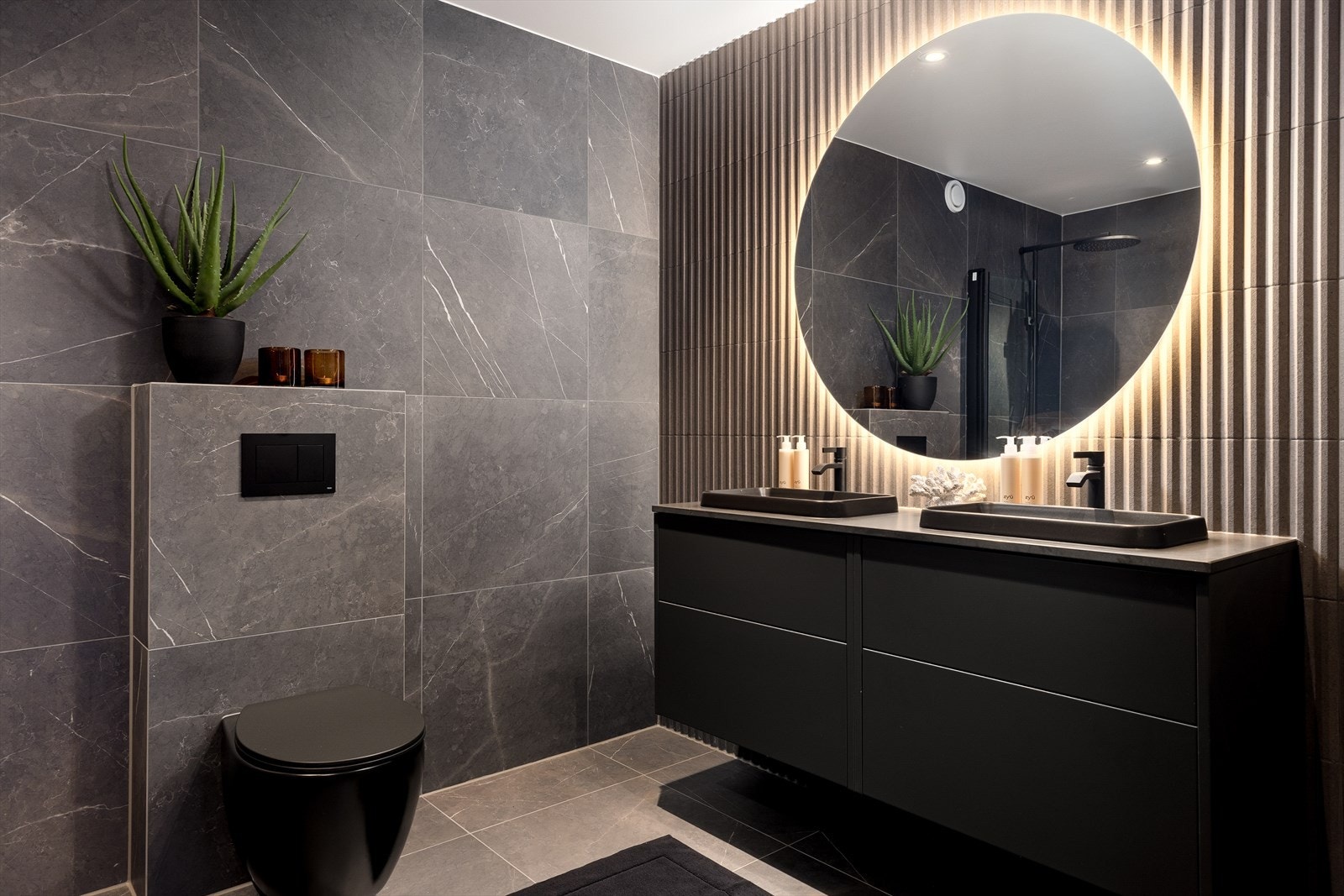 Lekkert flislagt bad med kontrastvegg. Skuffeseksjon med dobbelservant og speil med integrert belysning. Vegghengt wc. Galleribilde