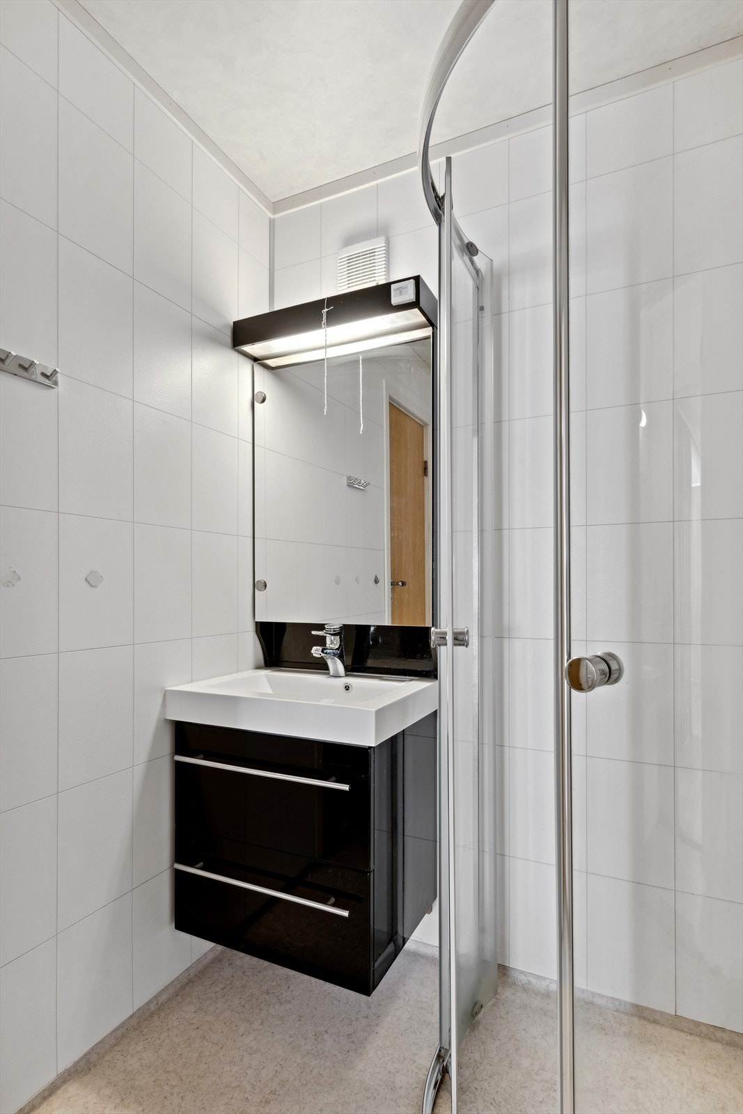 Rommet består av vinylbelegg med varmekabel, baderomsplater i dusjrom og folierte plater på wc. Innredet med dusjdører med sokkellist og en liten baderomsinnredning med skuffer og heldekkende servant. Galleribilde