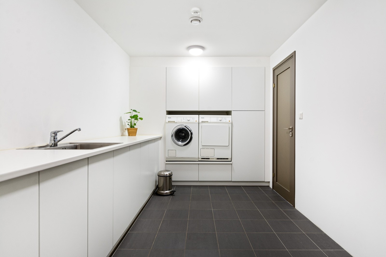 Vaskerommet har mørke gulvfliser, lysmalte vegger, varme i gulvet og avtrekk via ventilasjonsanlegget. Skapene omkranser en sokkel hvor opplegg for vaskemaskin og tørketrommel er plassert i god arbeidshøyde. Galleribilde