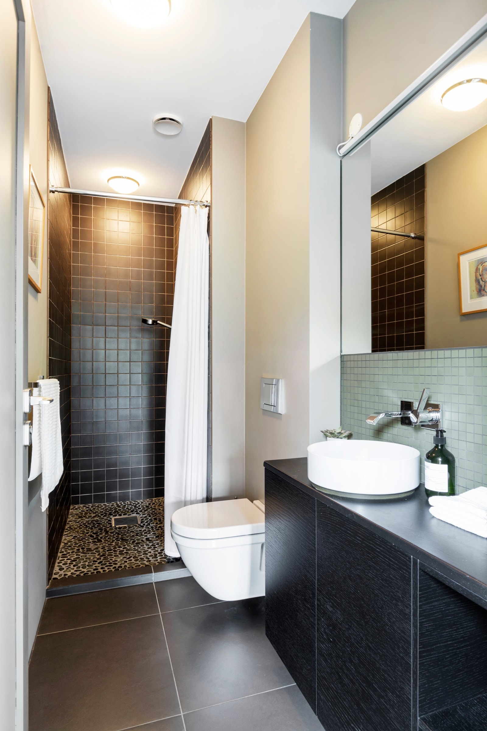 Bad med adkomst fra gangen i andre etasje er holdt i samme stil som badet i underetasjen med mørke gulvfliser i 60 x 60 cm og rullestein i dusjsonen. Det er speil, ledbelysning, integrert blandebatteri, vegghengt toalett, og gulvvarme. Galleribilde