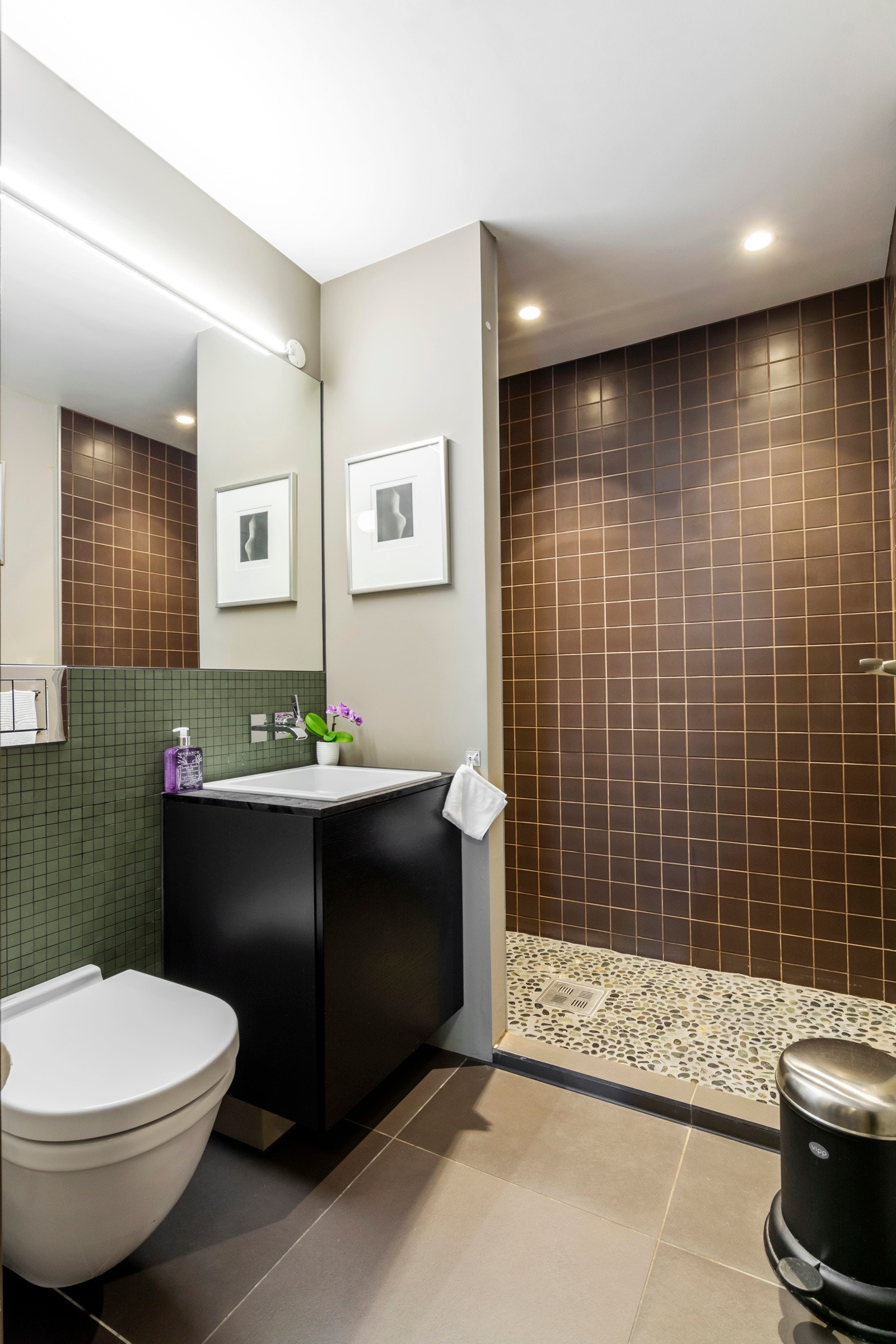 Badet i underetasjen har mørke gulvfliser i 60 x 60 cm, lekkert kombinert med rullestein i dusjsonen. Veggene har malte flater og kledelige mosaikkfliser. Det er speil, led-belysning, integrert blandebatteri, samt hvit servant nedfelt i mørk innredning. Galleribilde