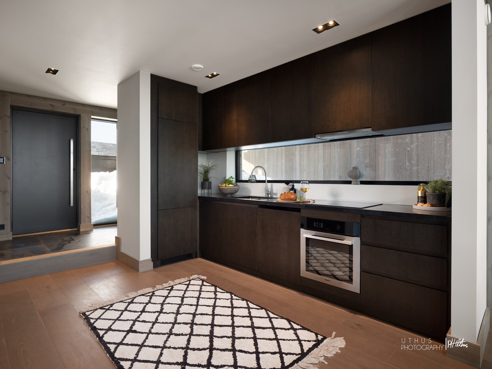 Mørkfarget eik med enkle slette fronter, med gripelister og benkeplate i Quartz type Piatra Gray. Dempebeslag på alle skuffer og skap. Galleribilde