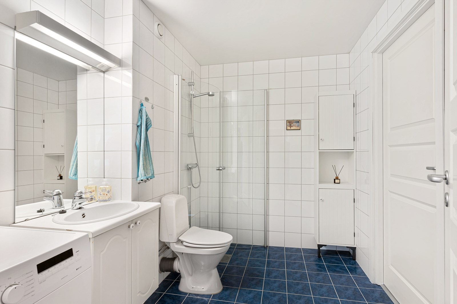 Badet har hvite veggfliser, mørkeblå gulvfliser med gulvvarme og opplegg for vaskemaskin. Galleribilde