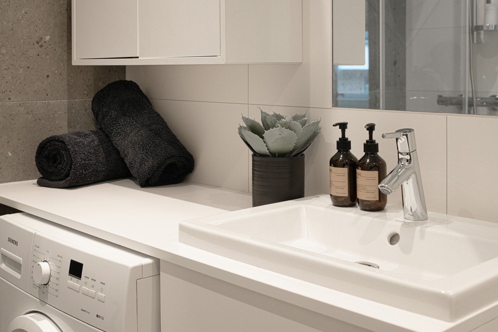 Tidløs baderomsinnredning med hvite fronter og benkeplate med nedfelt servant, speil og vegghengt skap over. Praktisk med plass og opplegg for vaskemaskin under benkeplaten. Galleribilde