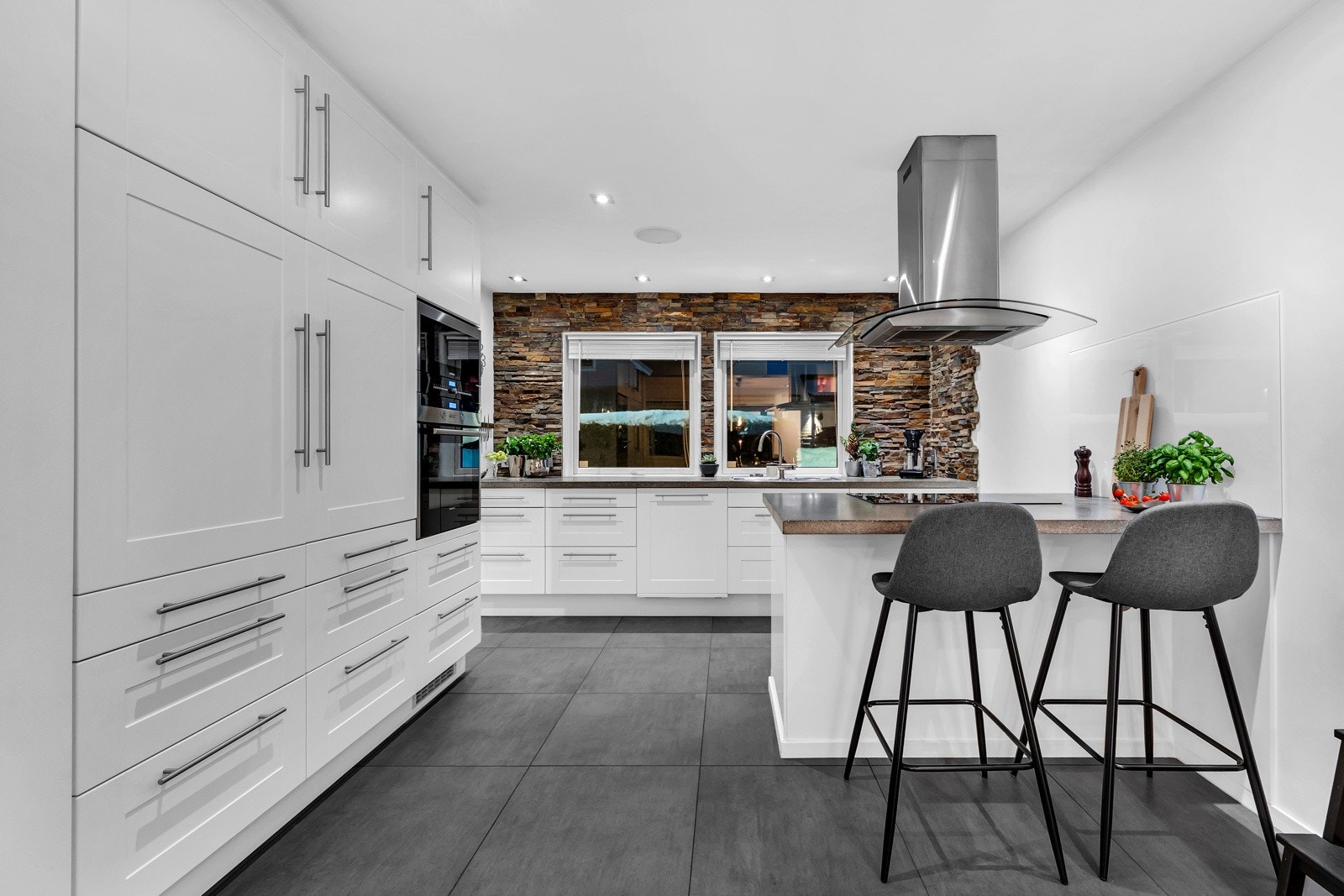 Kjøkkenet ligger i åpen løsning til stuen/spisestuen. Rommet er holdt i samme moderne farger som stuen og flisene i entréen er videreført inn på kjøkkenet. Kjøkkenet har en kjempe stilig og unik steinbelagt vegg, som er prikken over i-en i rommet. Galleribilde