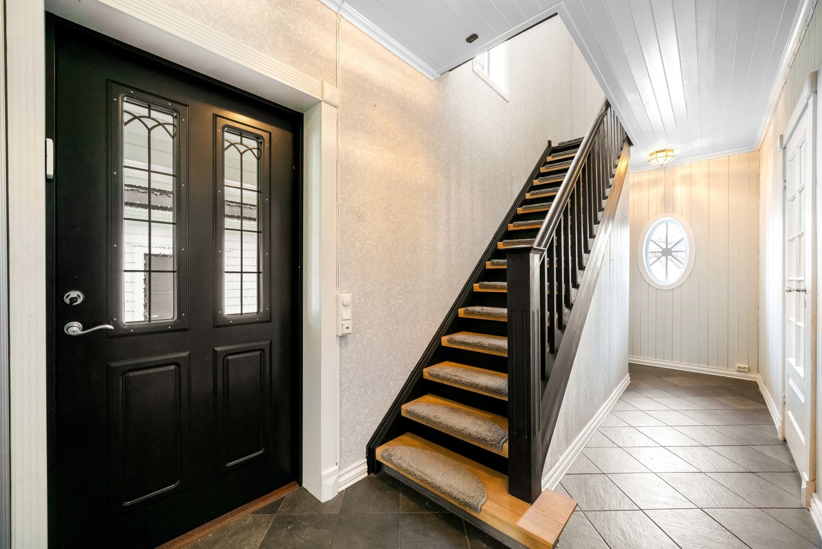 Du ønskes velkommen inn i en delikat entrè utstyrt med skyvedørsgarderobe i speil. Den originale trappen til 2.etasje har eiketrinn og sortmalt rekkverk. Gulvet er flislagt med grå fliser, og veggene er utført med lysmalt panel samt tapet under trappen. Galleribilde
