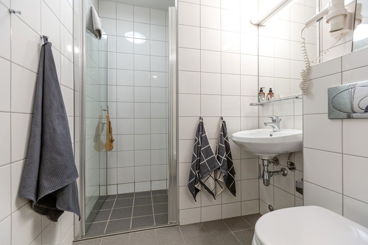 Romslig bad med dusjnisje, hvit porselensservant, vegghengt speil med lyslist over og vegghengt toalett Galleribilde