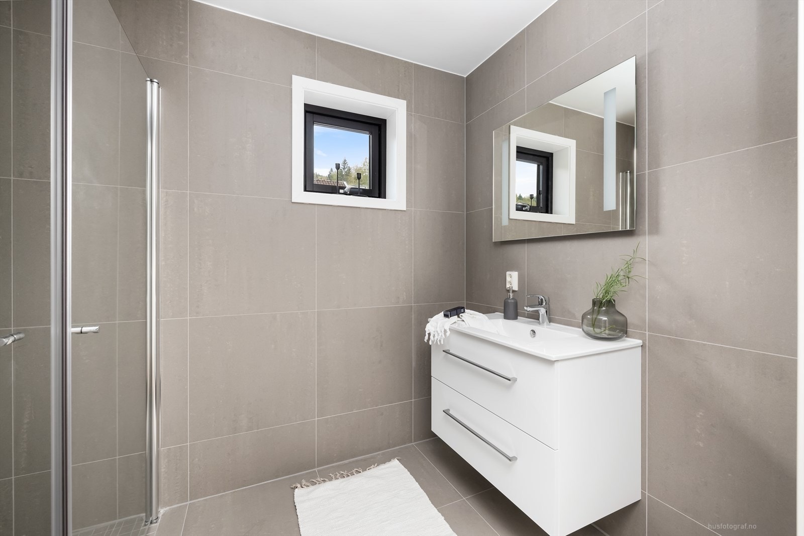 Bad har sanitærutstyr bestående av veggmontert klosett, dusjhjørne, servant m/baderomsinnreding og overspeil. Galleribilde