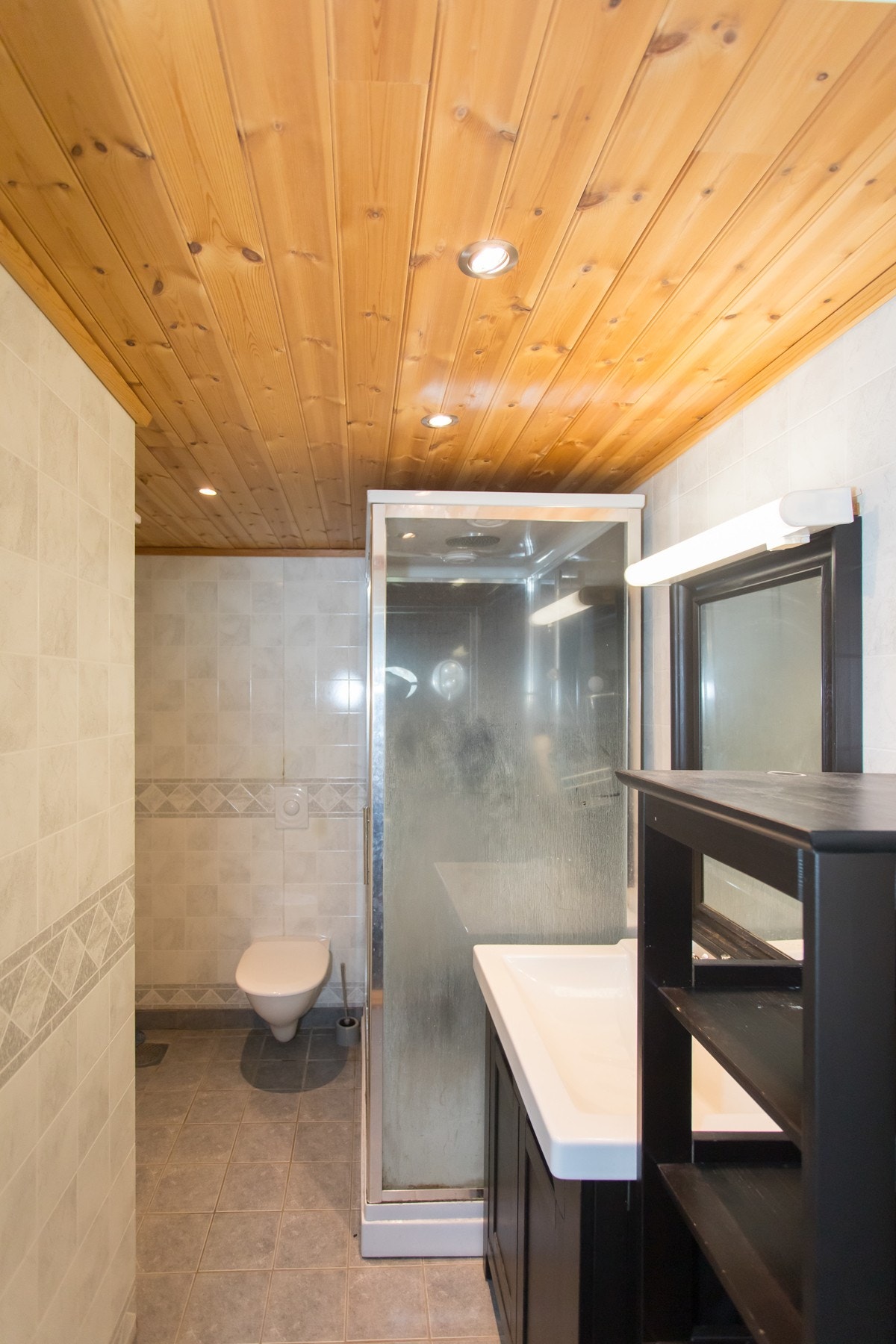 Bad i hybeldelen. Badet har behov for renovering. Her er det innredet med fliser med varmekabler på gulv og baderomsplater på vegg. Innredet med dusjkabinett, toalett og baderomsinnredning. Galleribilde