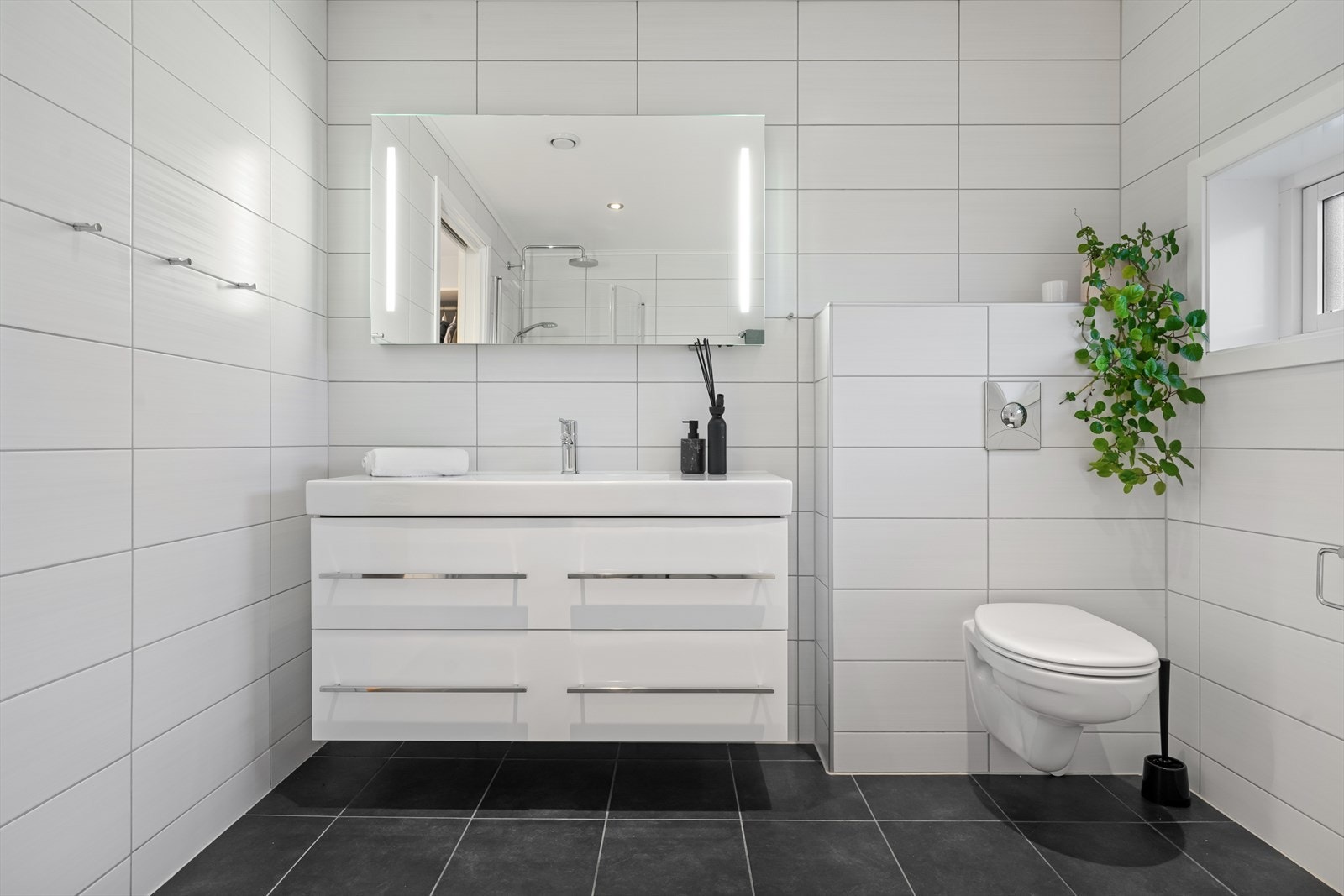 Badet er stilrent med sorte gulvfliser og hvite veggfliser for en fin kontrast. Videre har badet gulvvarme, downlights og vegghengt toalett Galleribilde
