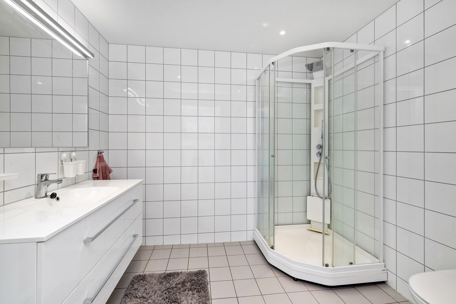 Lyst flislagt bad med dusjkabinett, vegghengt toalett, samt baderomsinnredning med porselenbenk/vask, speil med belysning. Galleribilde