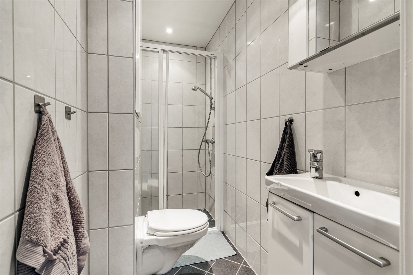 Badet inneholder vegghengt toalett, helstøpt servant, skap under servant med slette fronter og dusjdører i klart glass. Galleribilde