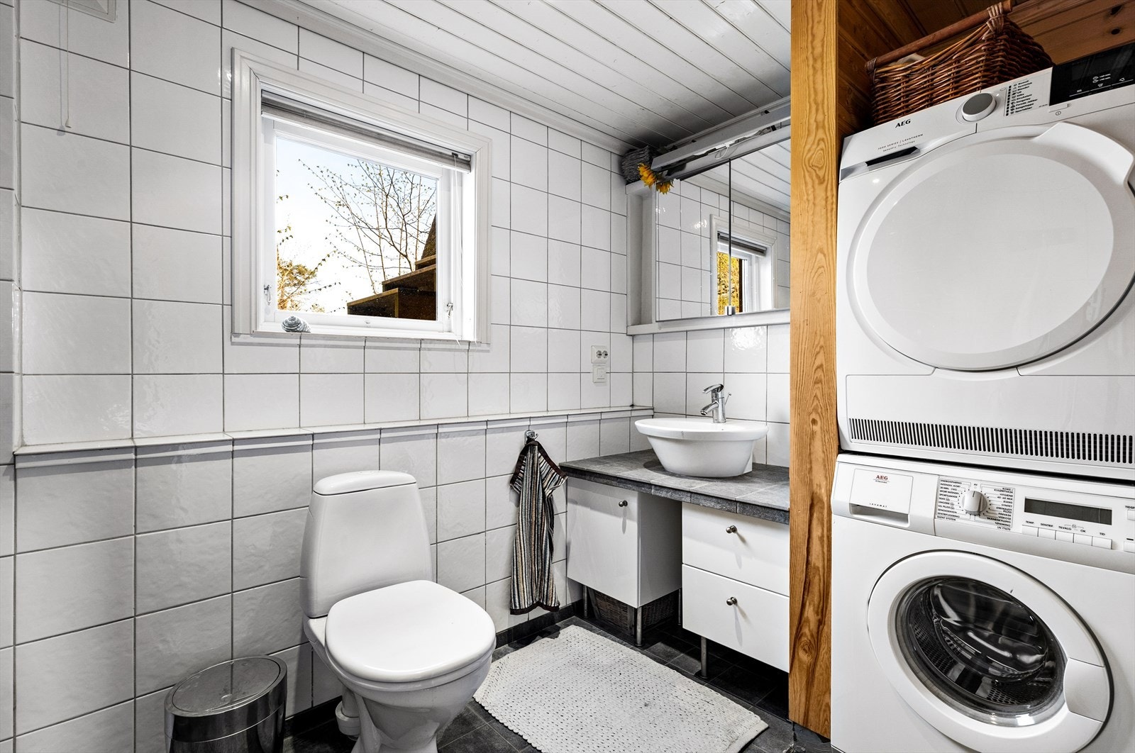 Bad/vaskerom innredet med toalett, servantskap, flislagt benkeplate og overmontert hvit keramisk enkel servant med ettgreps blandebatteri i rustfritt stål fra Grohe. Opplegg for vaskemaskin Galleribilde