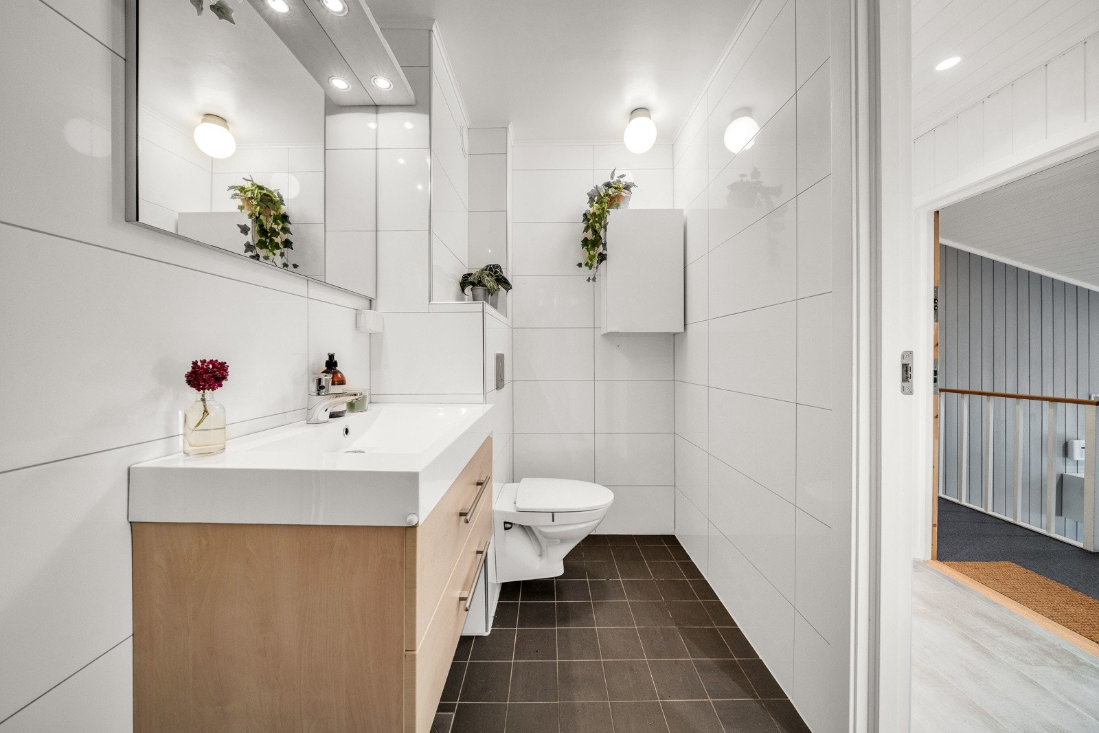 .. Videre inneholder badet servantskap med glatte fronter, og speil med lysarmatur over, veggmontert wc, dusjhjørne med svingbare glassvegger, og opplegg for vaskemaskin Galleribilde