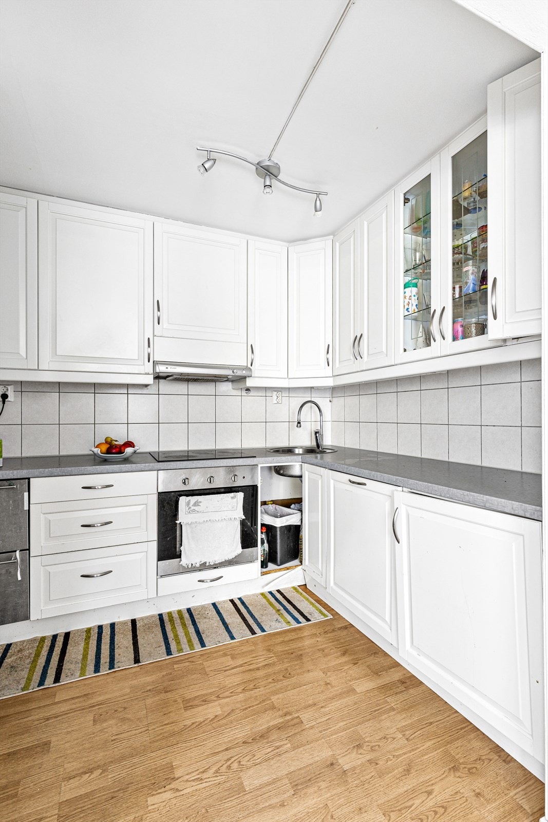 Kjøkkenet som ligger i delvis åpen løsning fra stuen består av hvit laminat skapinnredning med profilerte fronter, vitrineskap og nedfelt kum med ett-greps kran. Laminat benkeplate. Fliser mellom benkeplate og overskap. Mekanisk avtrekk Galleribilde