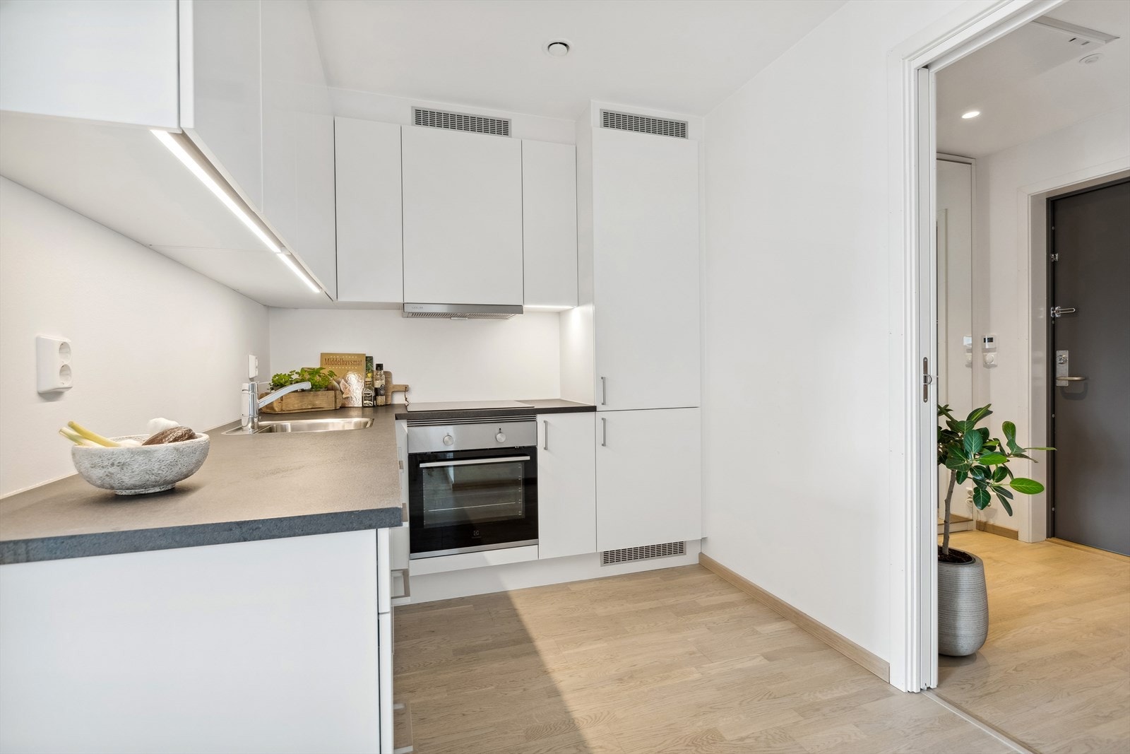 Tiltalende kjøkkeninnredning i hvit utførelse med laminat benkeplate. Overskapene går helt opp til himling. Innredningen er utstyrt med integrert koketopp, stekeovn, oppvaskmaskin og kjøleskap. Det er montert leddlys under overskap. Galleribilde