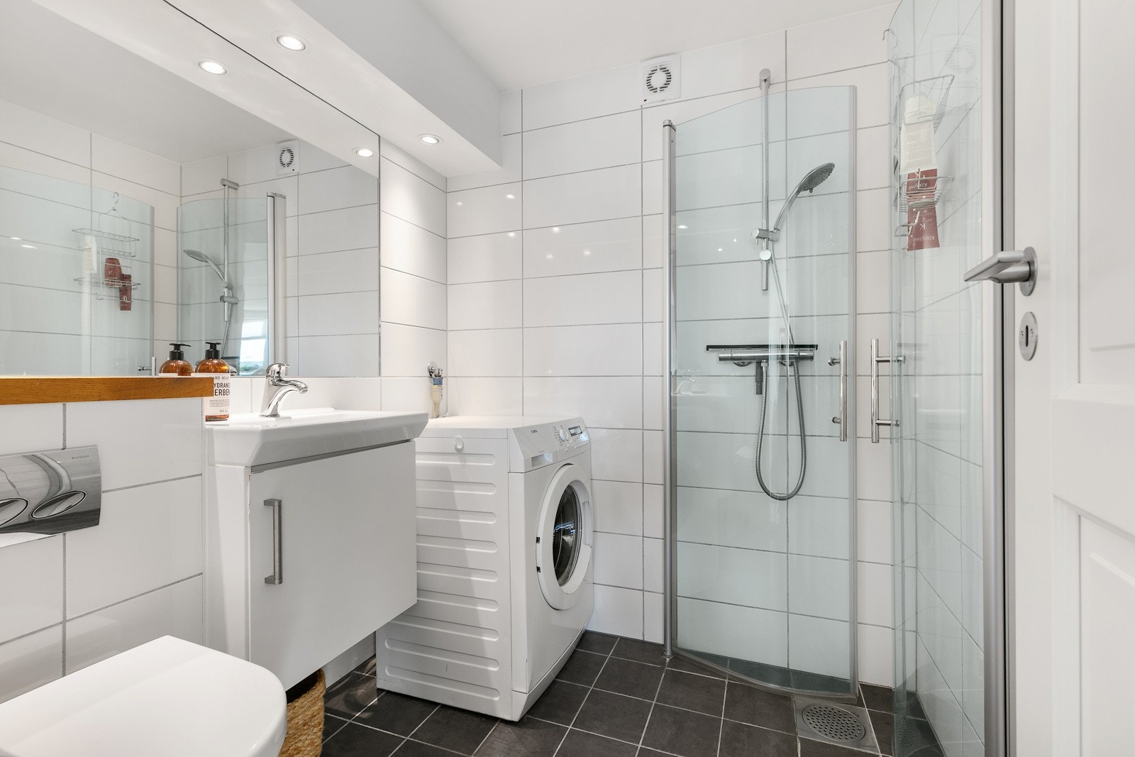 Badet i hybelen har mørkegrå fliser på gulv med gulvvarme, hvite fliser på veggene, downlights og opplegg for vaskemaskin. Galleribilde