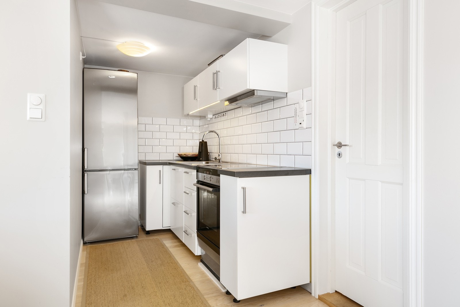 Kjøkkenet er plassert i egen nisje av rommet. Det har hvite, glatte fronter, mørk benkeplate i laminat med nedfelt vaskekum og hvite metrofliser over benken. Innredningen har frittstående komfyr/stekeovn, ventilator og plass til kjøleskap. Galleribilde