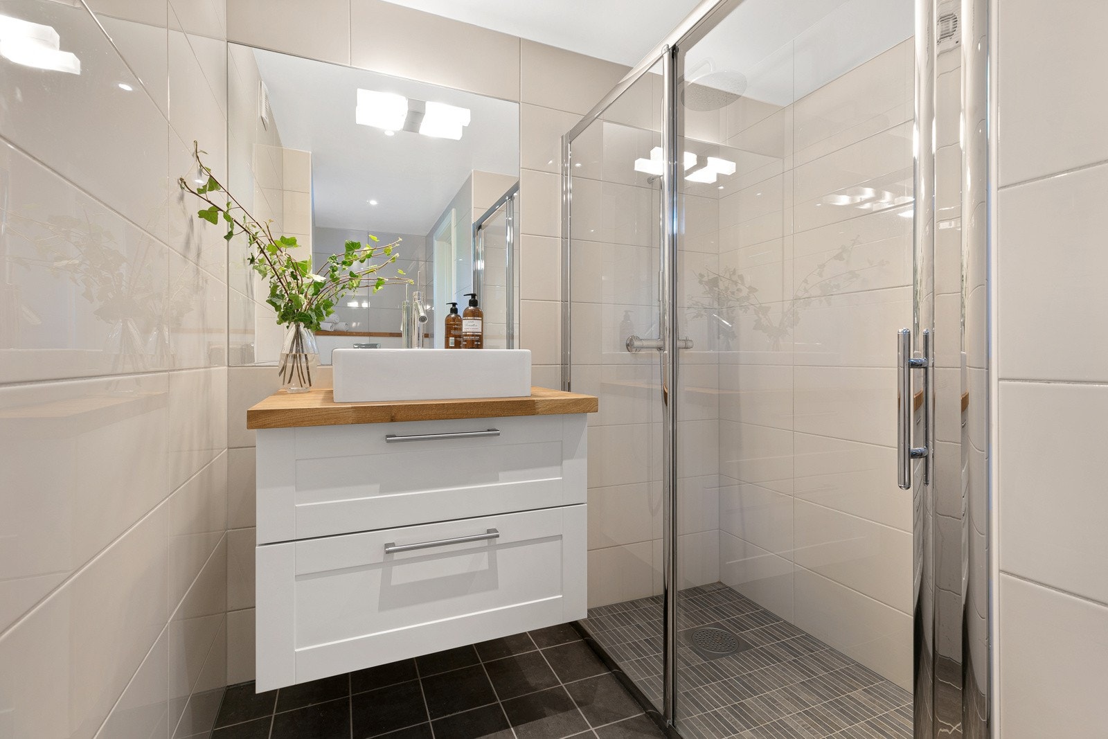 Badet i underetasjens hoveddel har mørke fliser på gulv, lyse, glossy veggfliser, gulvvarme, downlights og speilbelysning. Innredningen består av en toppmontert servant på hvit skuffeseksjon med eikeplate, speil, veggmontert toalett og et dusjhjørne. Galleribilde