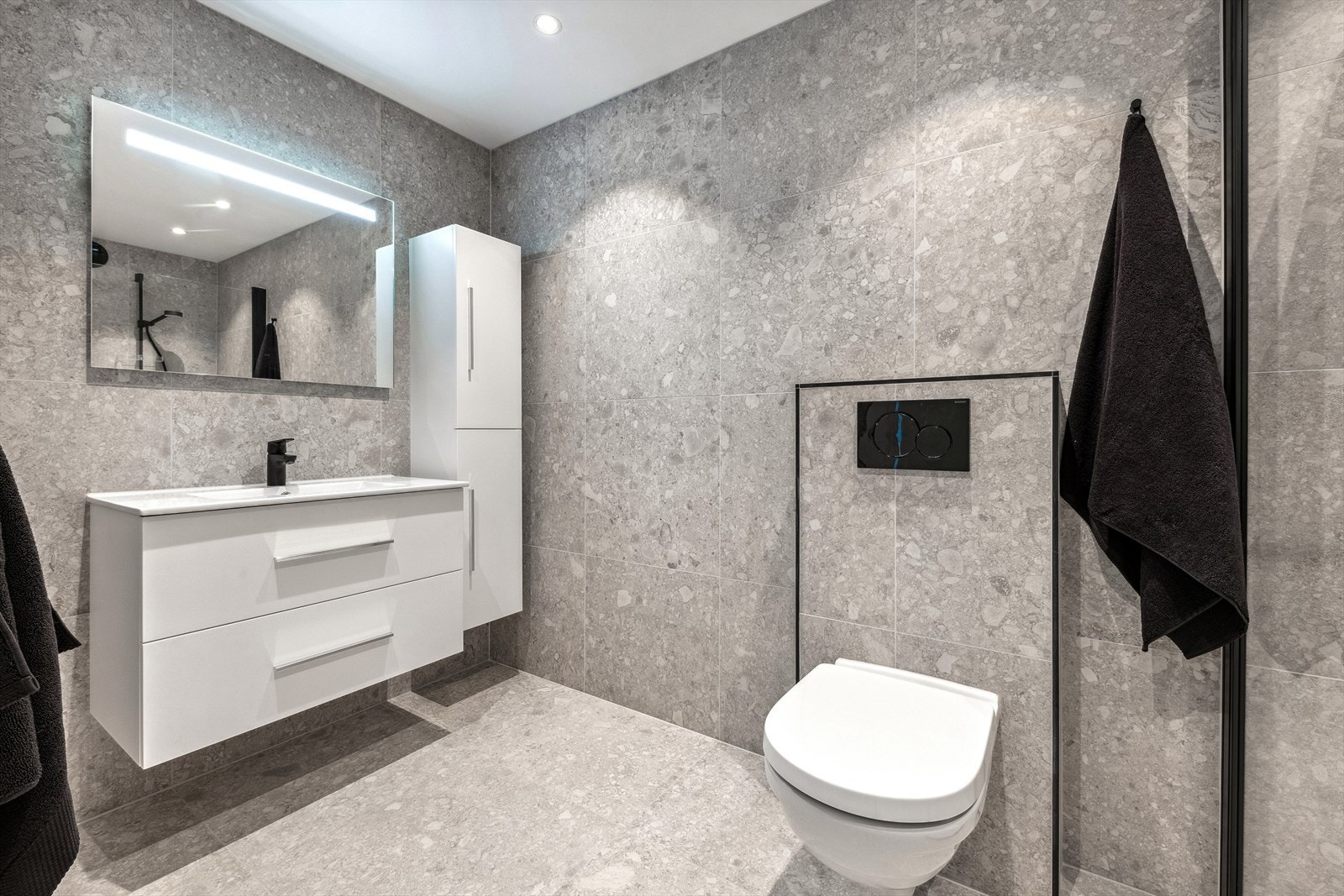 Lys baderomsinnredning med servant, speil med integrert belysning, høyskap, veggmontert toalett, dusjhjørne med innfellbare glassdører og opplegg for vaskemaskin. Galleribilde