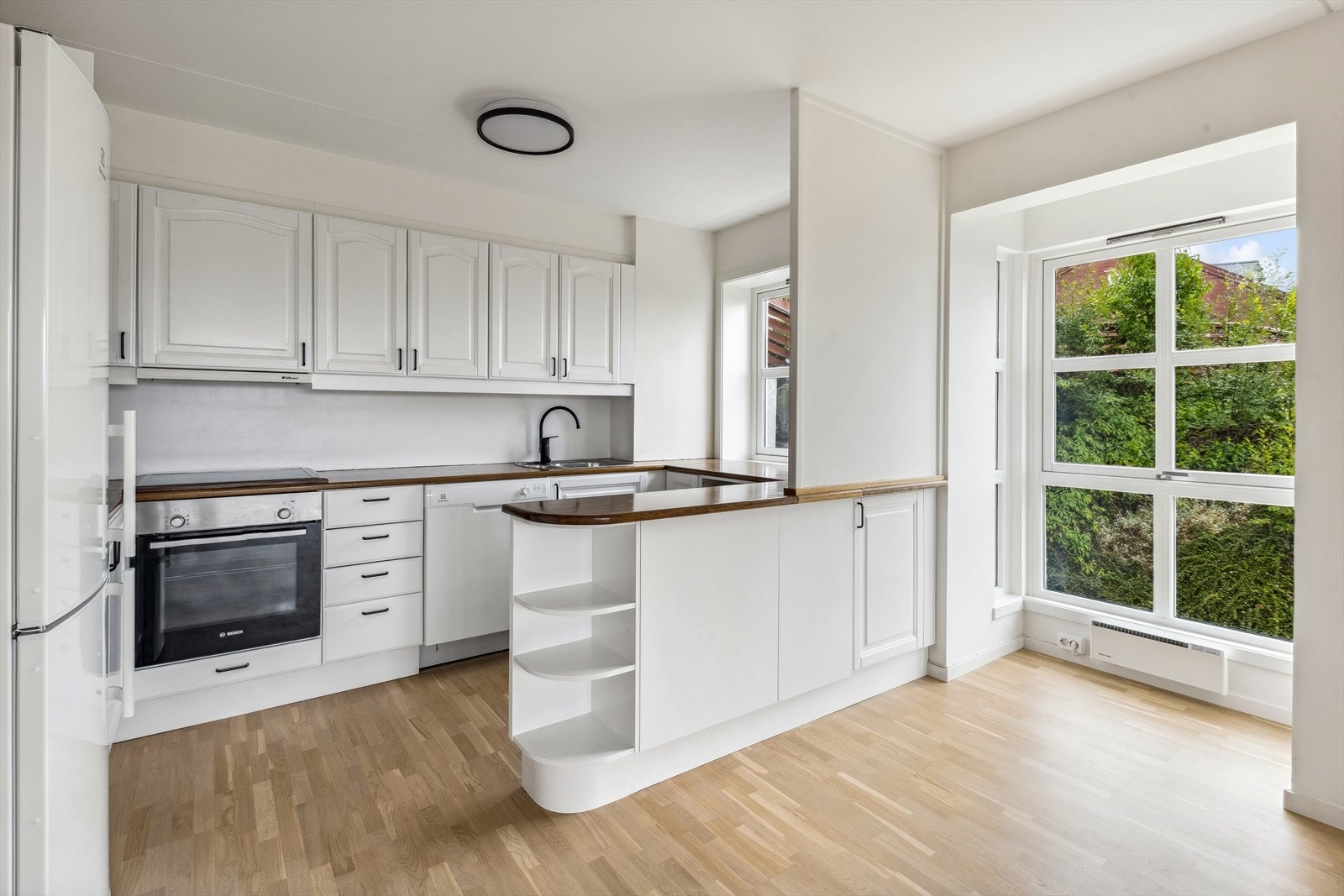 Lys kjøkkeninnredning med profilerte eikefronter, heltre benkeplate i eik, oppvaskkum med ett-greps blandebatteri og ventilator over kokesonen. Galleribilde