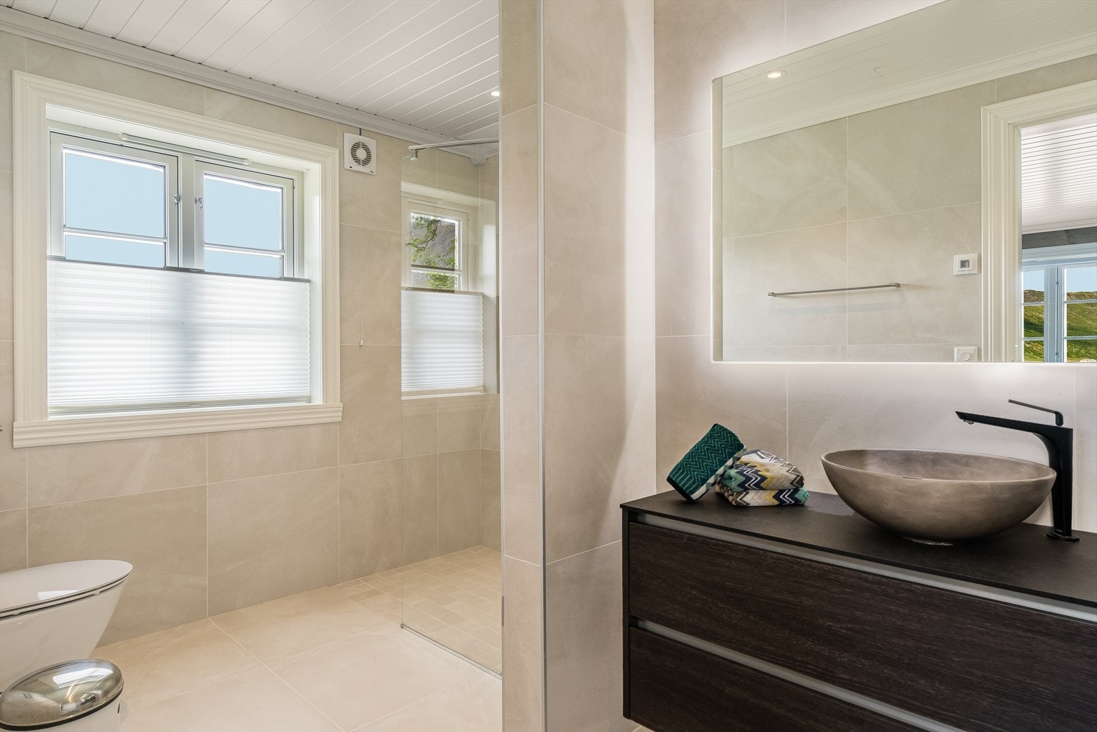 Pent helfliset bad med topp montert servant, veggmontert wc med innebygd sisterne, synlig lekkasjepunkt og dusjsone med glassvegg. Galleribilde