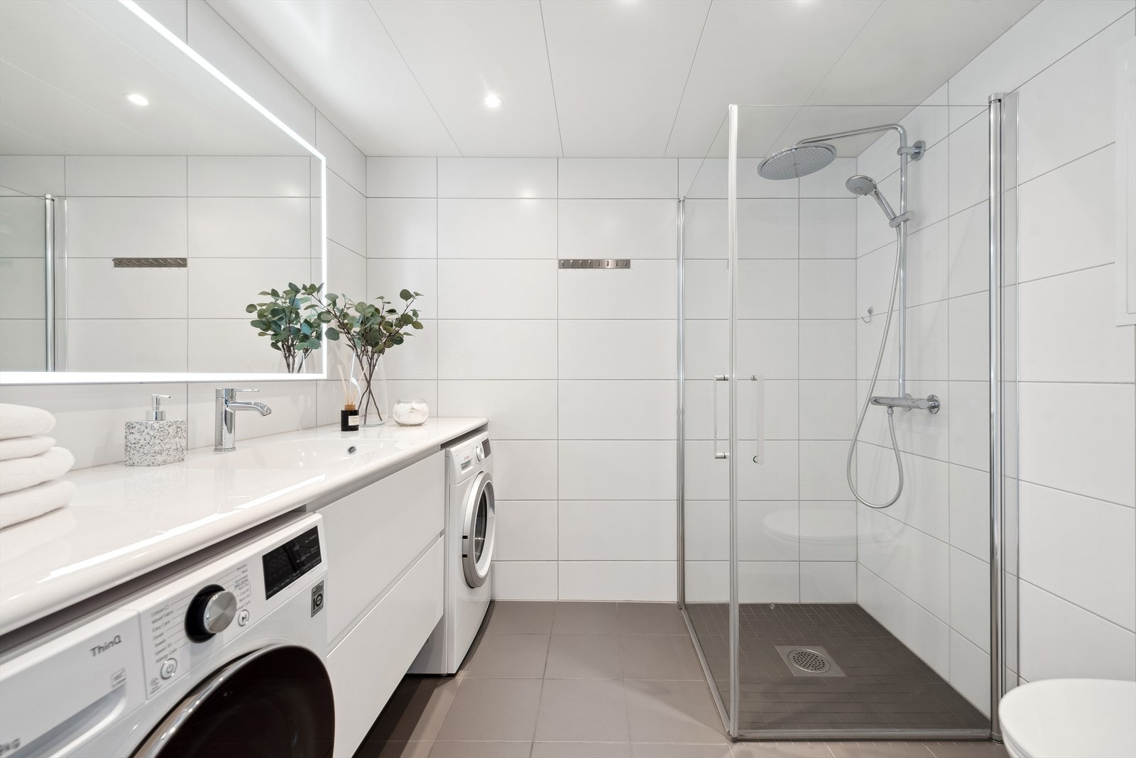 Lekkert flislagt bad med downlights i himling og varmekabler i gulv. Badet består av veggmonterte dusjvegger, dusj med termostat batteri, veggmontert klosett, servant med skuffer, speil med belysning og opplegg og plass for vaskemaskin og tørketrommel. Galleribilde