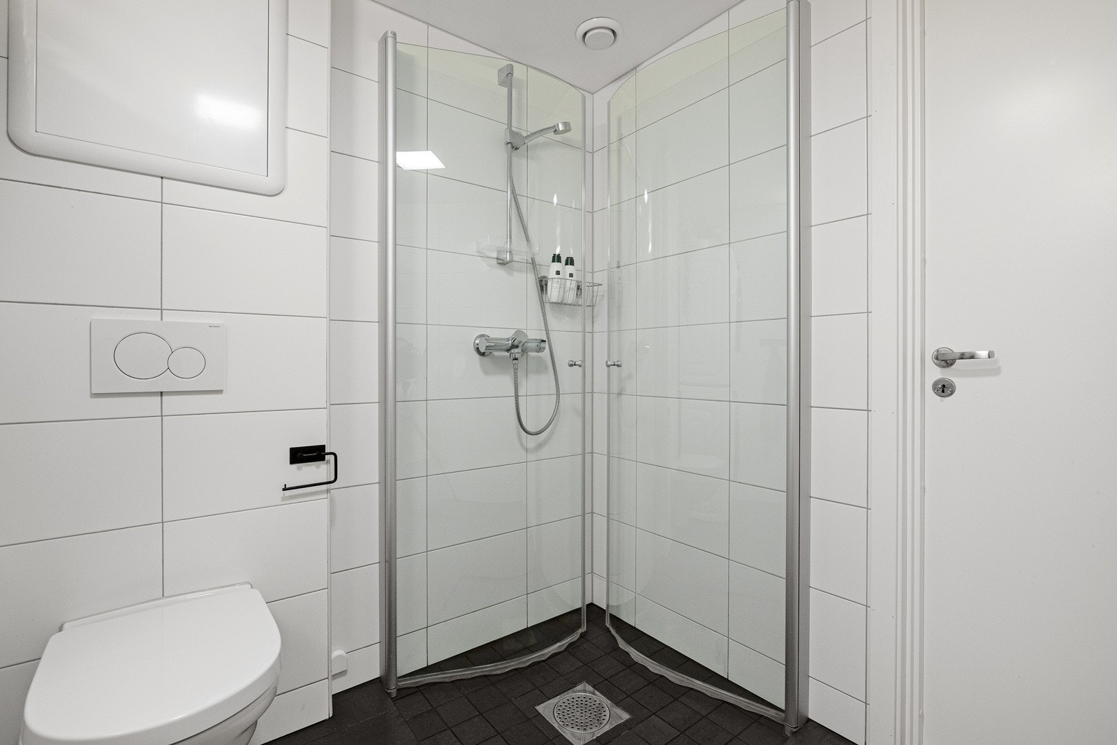 Dusjhjørne med glassdører og vegghengt dusjarmatur, vegghengt toalett og opplegg for vaskemaskin. Galleribilde