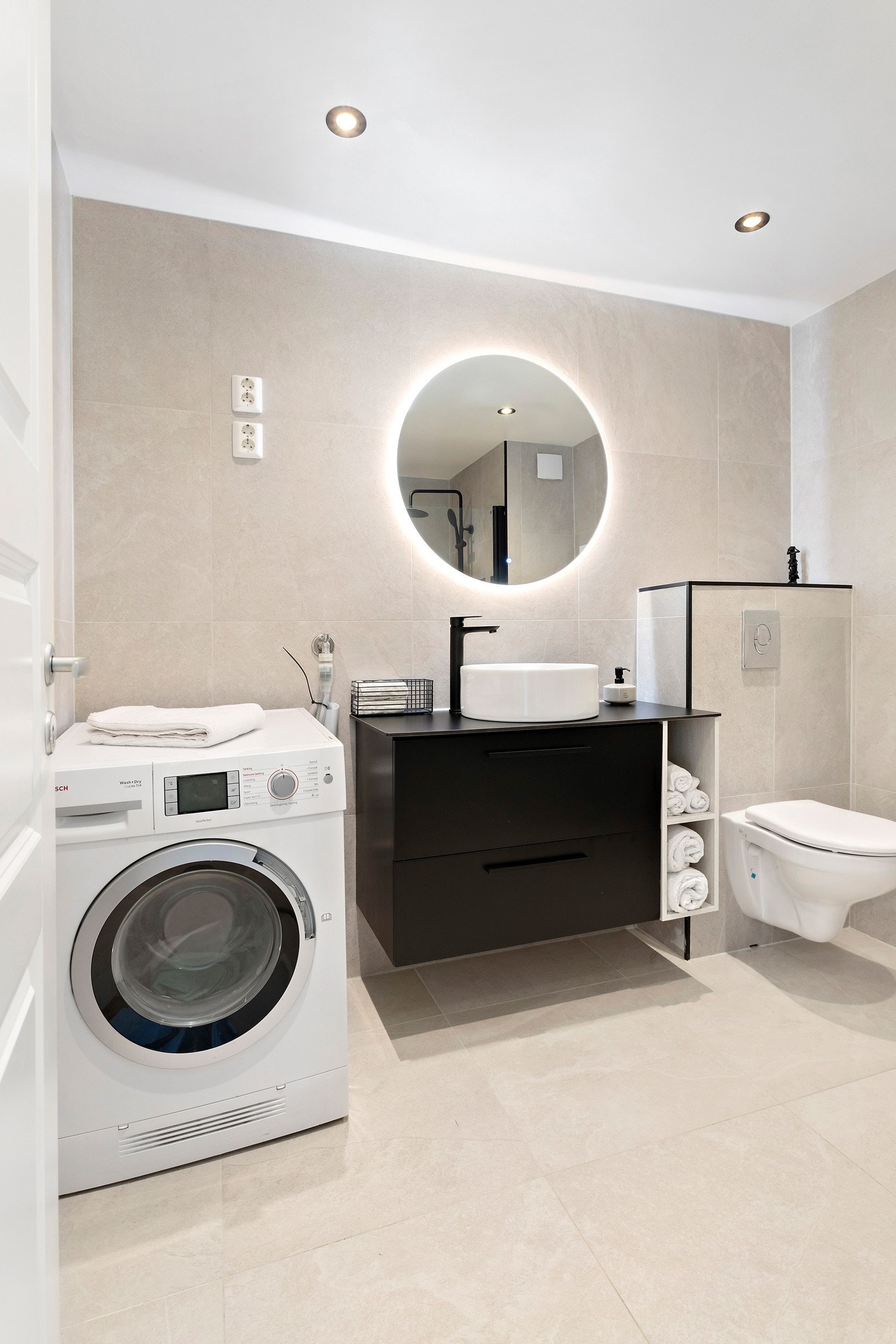 Pent baderom med store fliser på vegg og gulv i en lin tone med svarte detaljer på servant, sisterne og dusj. Galleribilde