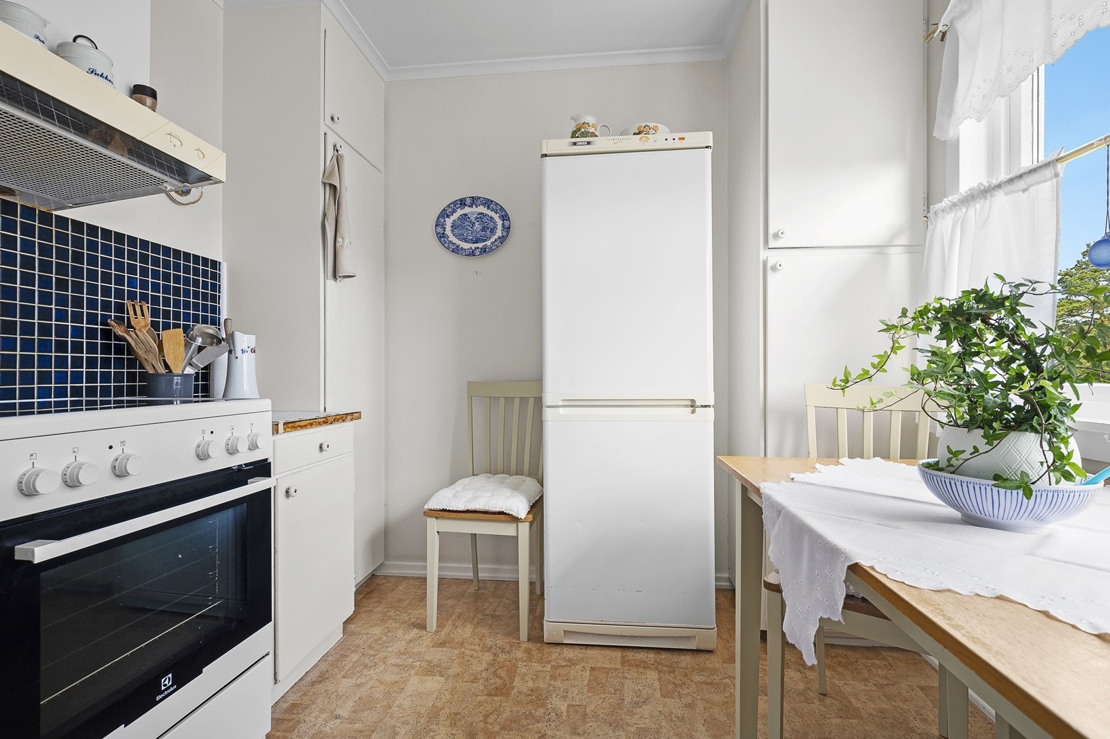 Kjøkken med eldre innredning som er hvitmalt, plassbygde skap og spiseplass. Galleribilde